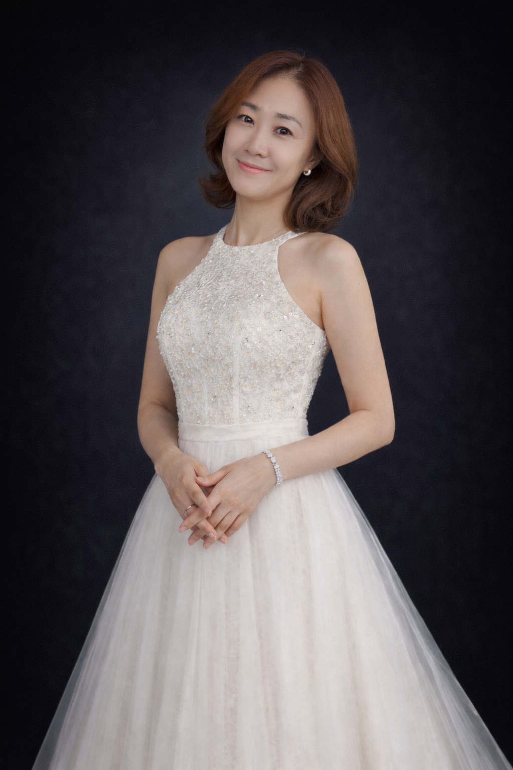 A woman in a white wedding dress with lace details on the bodice, smiling and holding her hands together, standing against a dark background.