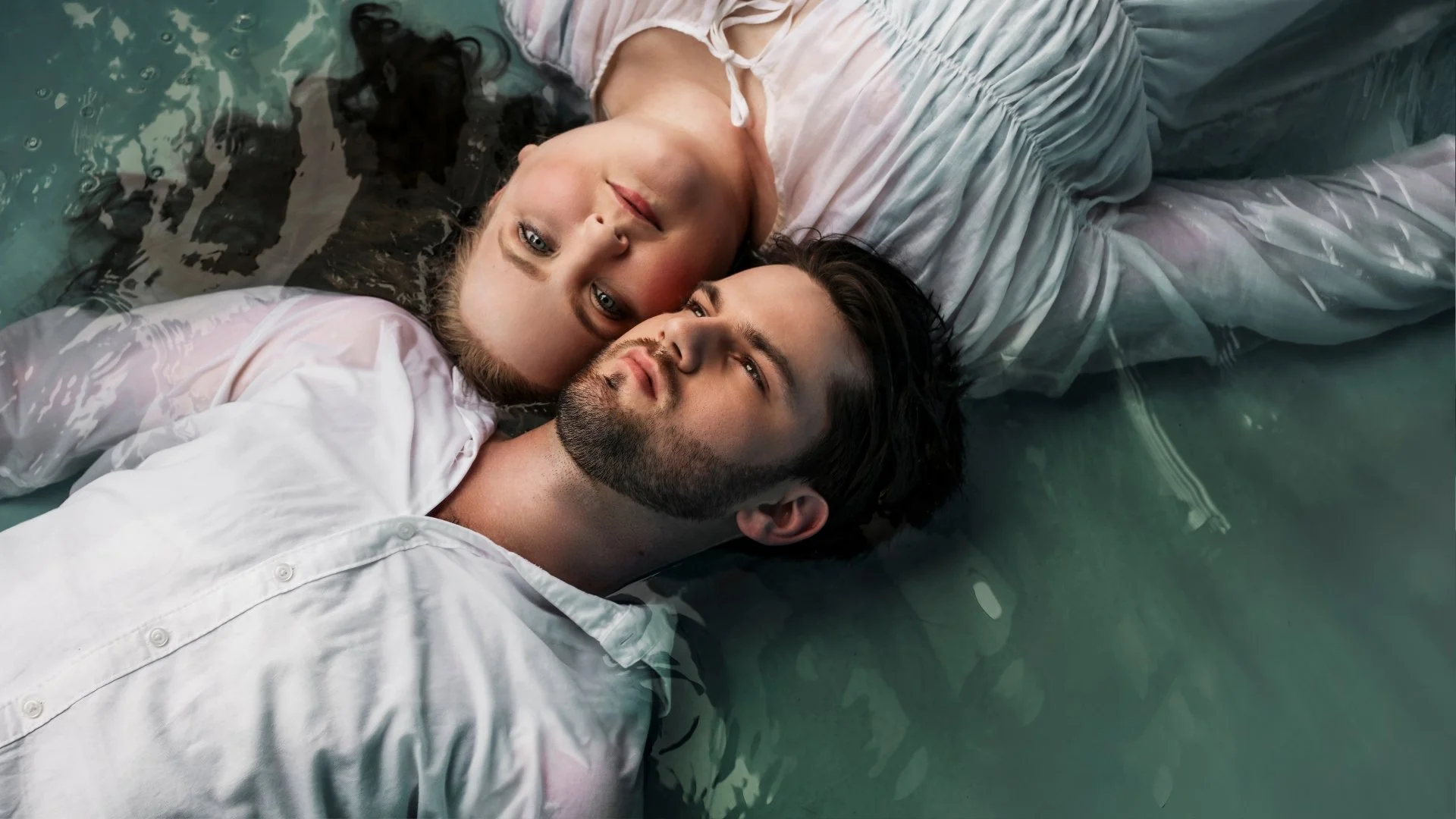 A man and woman are lying in water, their heads close together, with the woman's head resting on the man's shoulder. Both are looking upward, and they are dressed in white clothing.