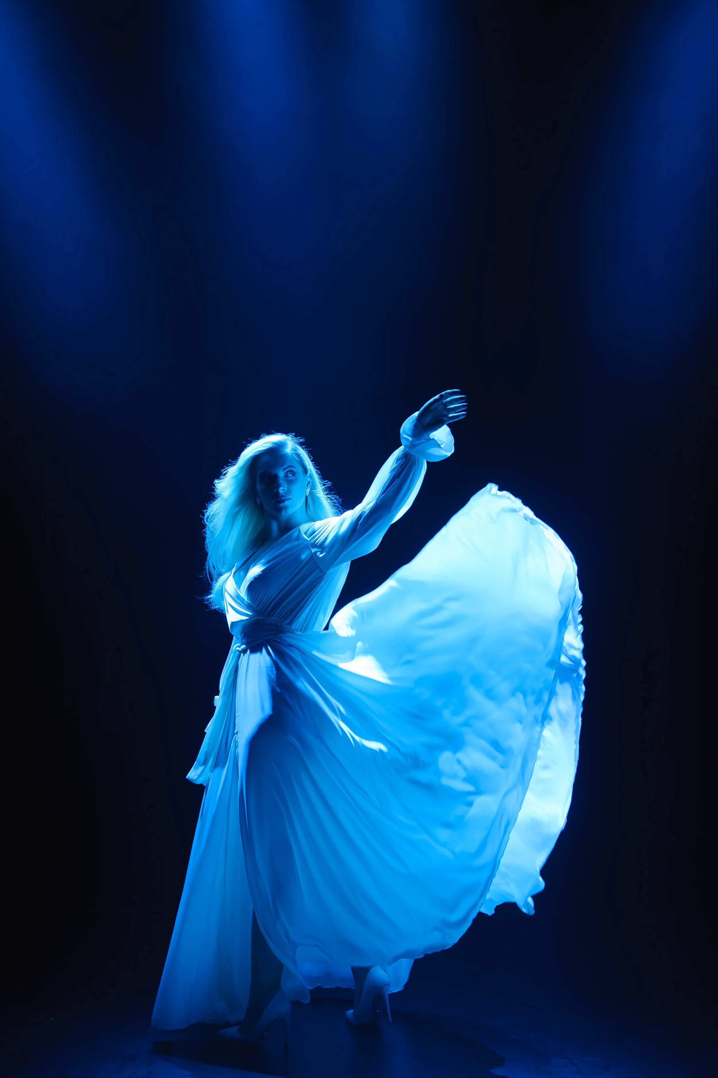 A woman in a flowing white dress performing a dance or dramatic pose on stage, illuminated by blue lighting.