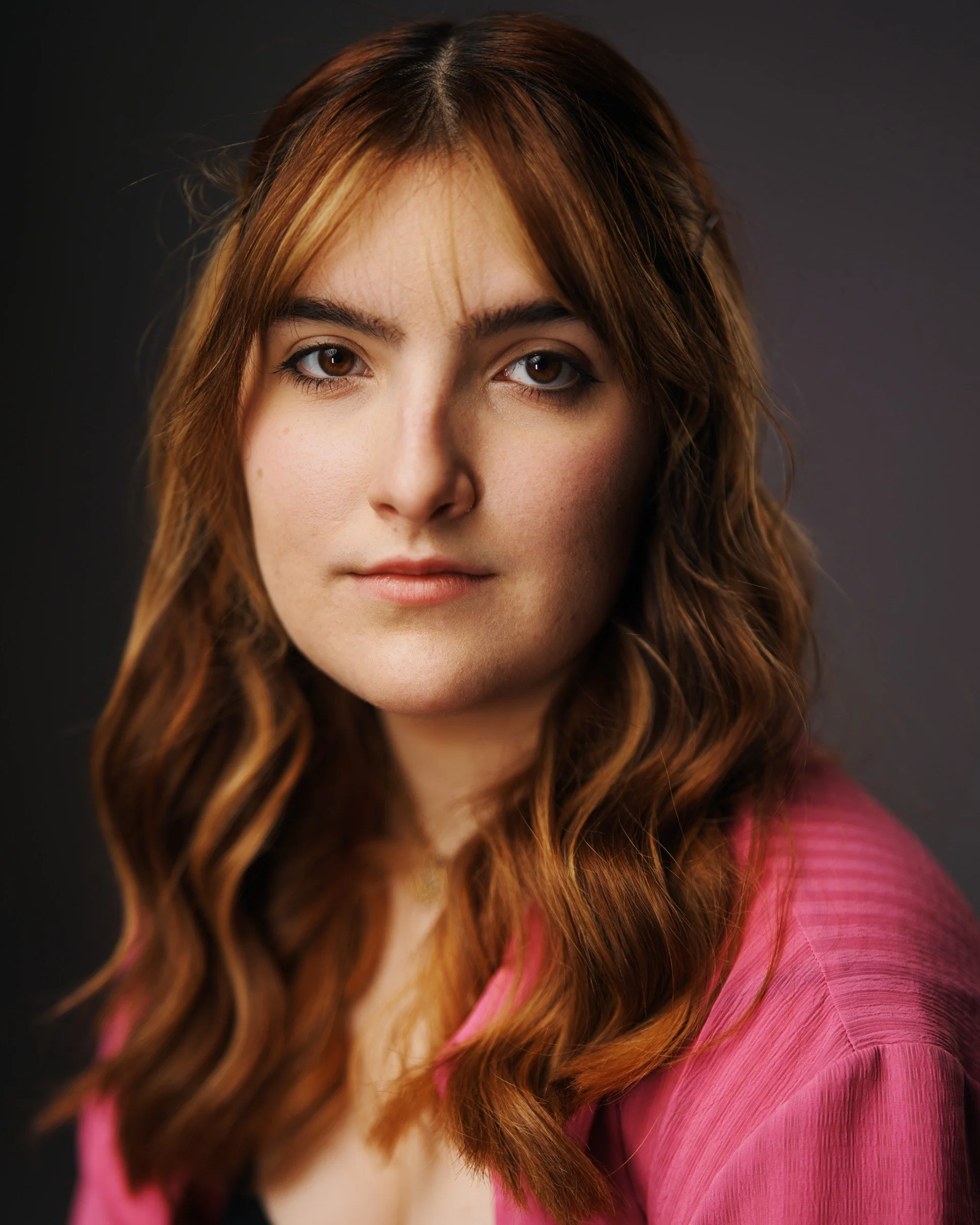 Close-up portrait of a young woman with wavy auburn hair wearing a pink top, against a dark background.