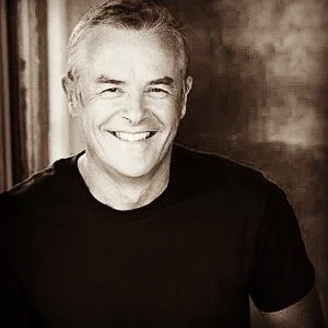 A smiling middle-aged man with short gray hair, wearing a black T-shirt, against a blurred dark background.