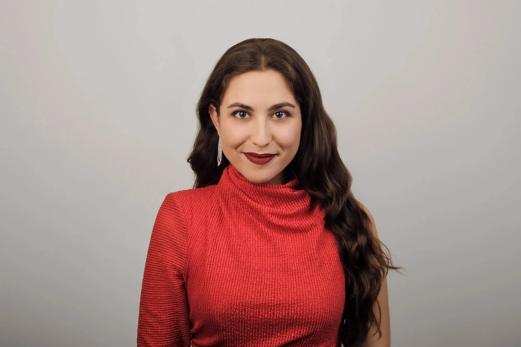 A woman with long dark hair wearing a red textured top and silver earrings, smiling in front of a plain light gray background.