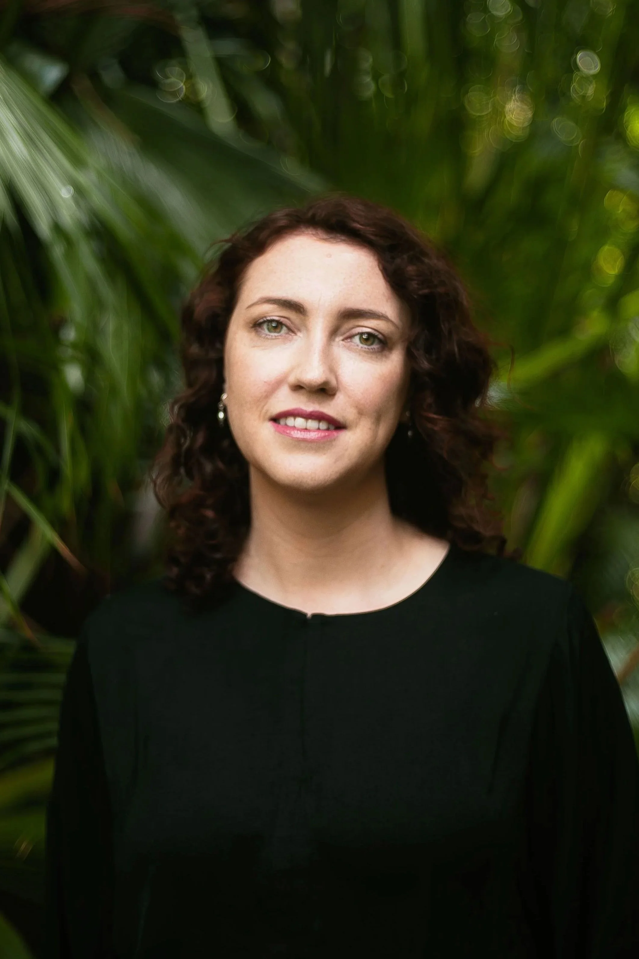 A woman with curly brown hair and light skin, wearing a black top and earrings, standing in front of green tropical foliage.