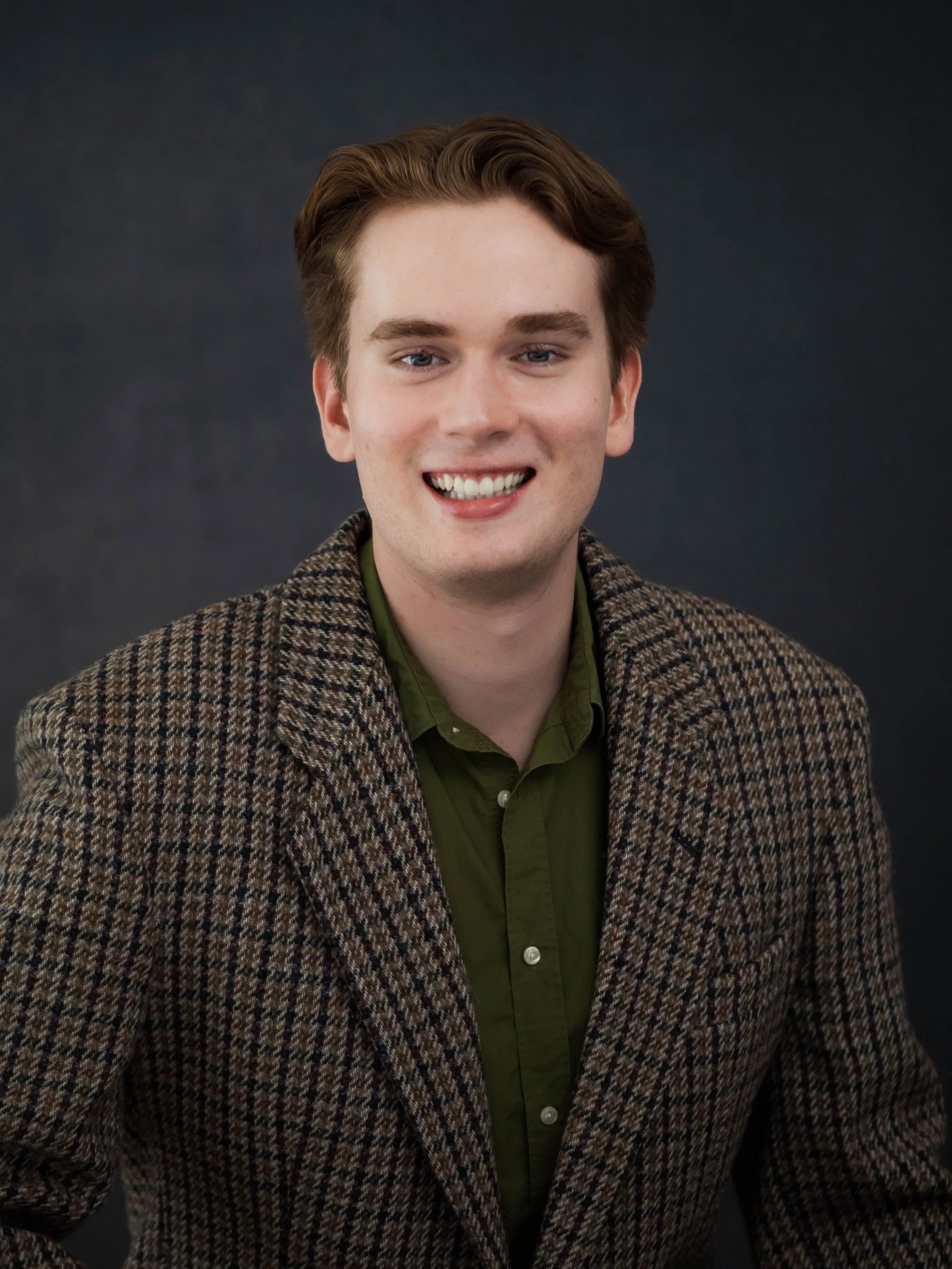Portrait of a young man with light skin, brown hair, wearing a green shirt and a brown checked blazer, smiling against a dark background.