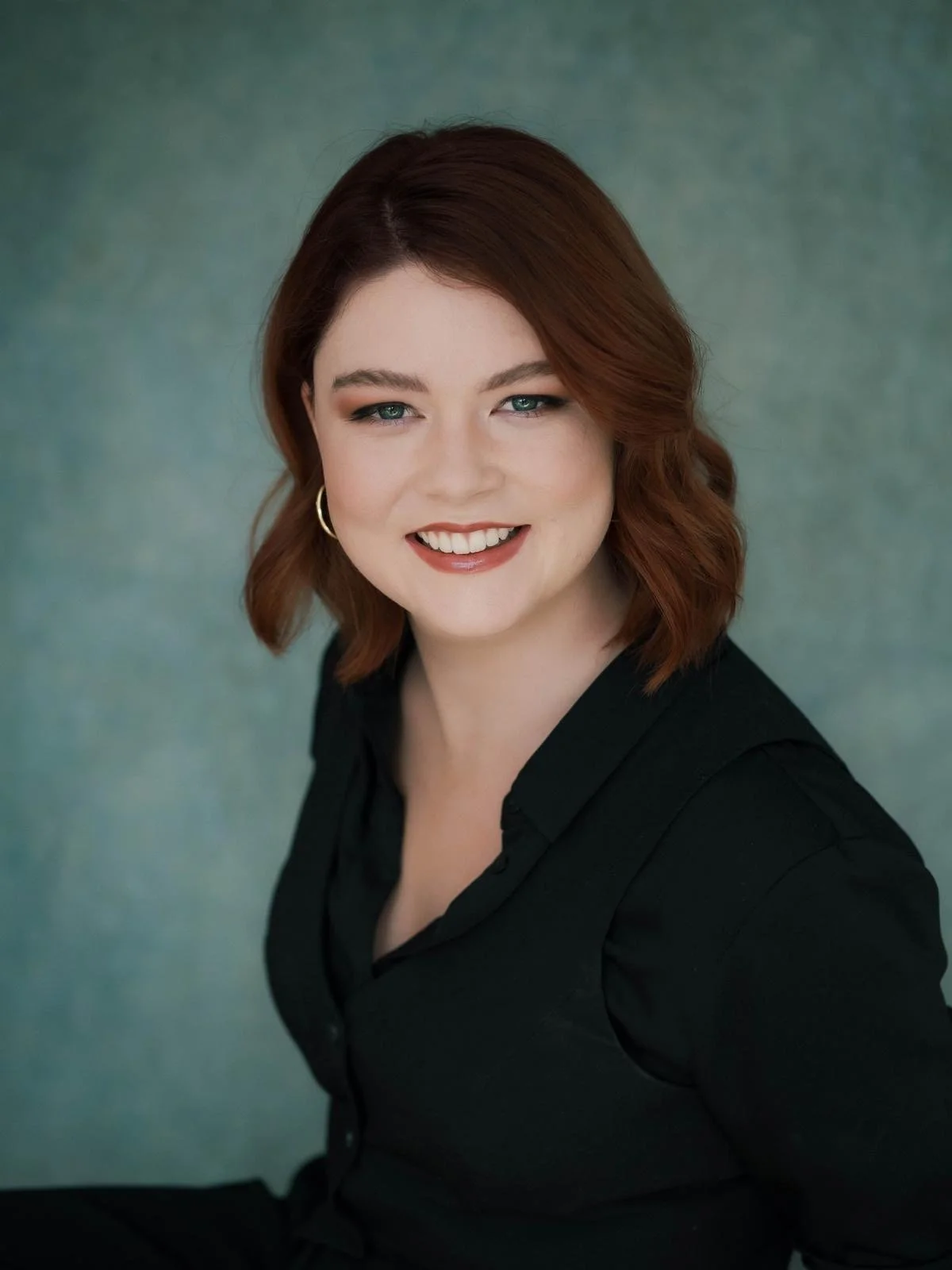 Portrait of a smiling woman with red hair, wearing a black top, and gold hoop earrings, against a textured green background.