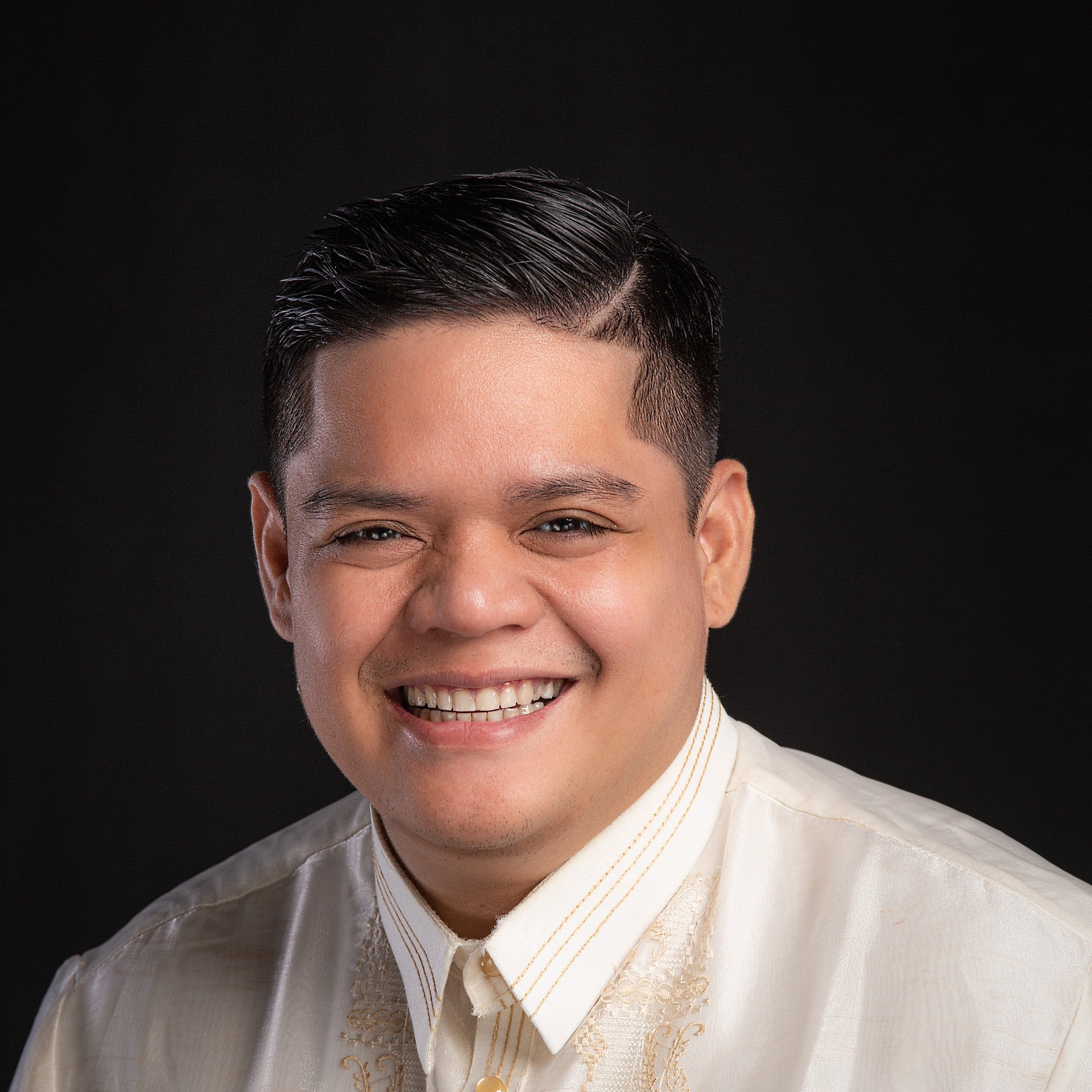 A smiling man with short black slicked-back hair, wearing a cream traditional Filipino Barong Tagalog, against a black background.