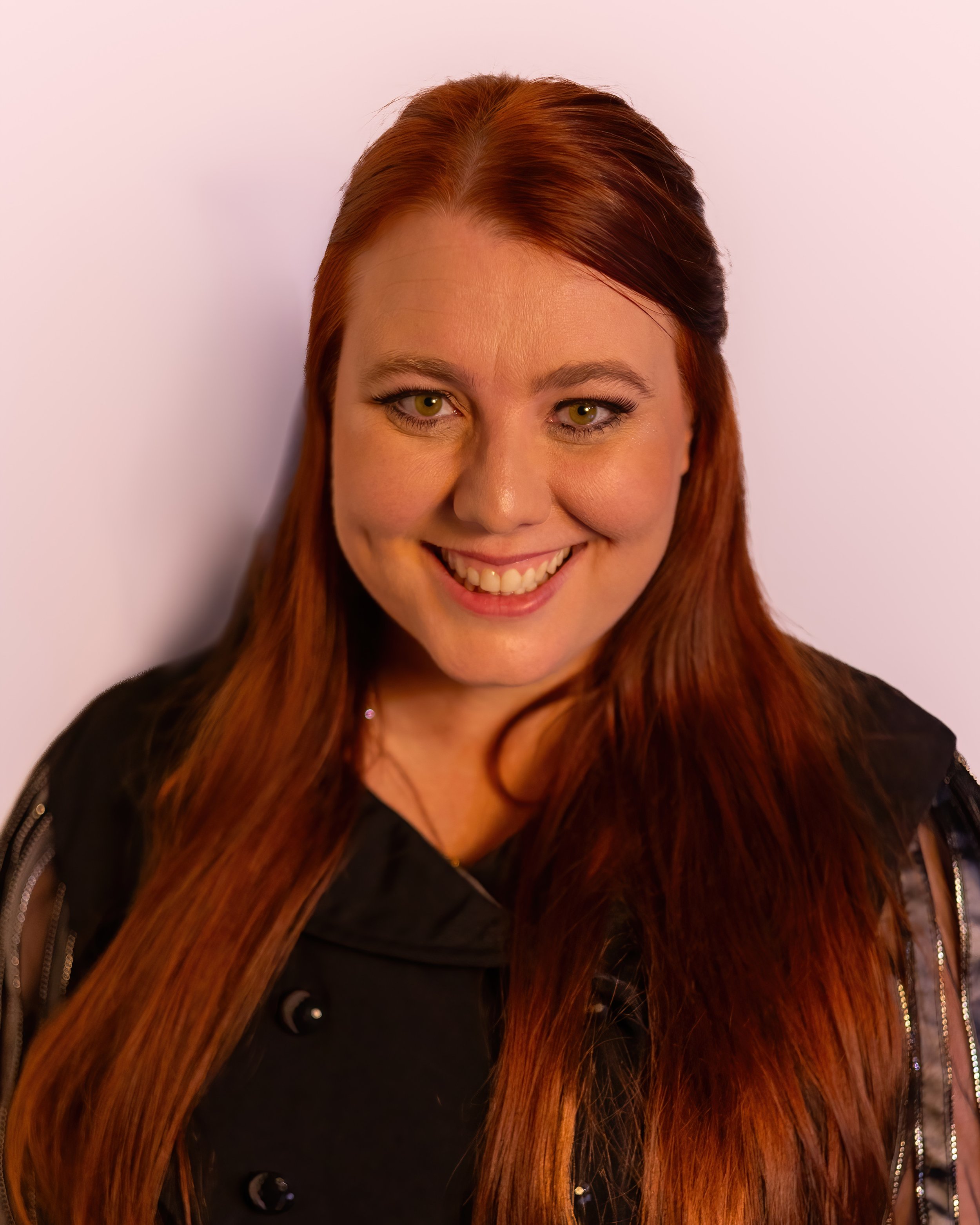 Portrait of a woman with long red hair, smiling, wearing a black jacket, against a plain white background.