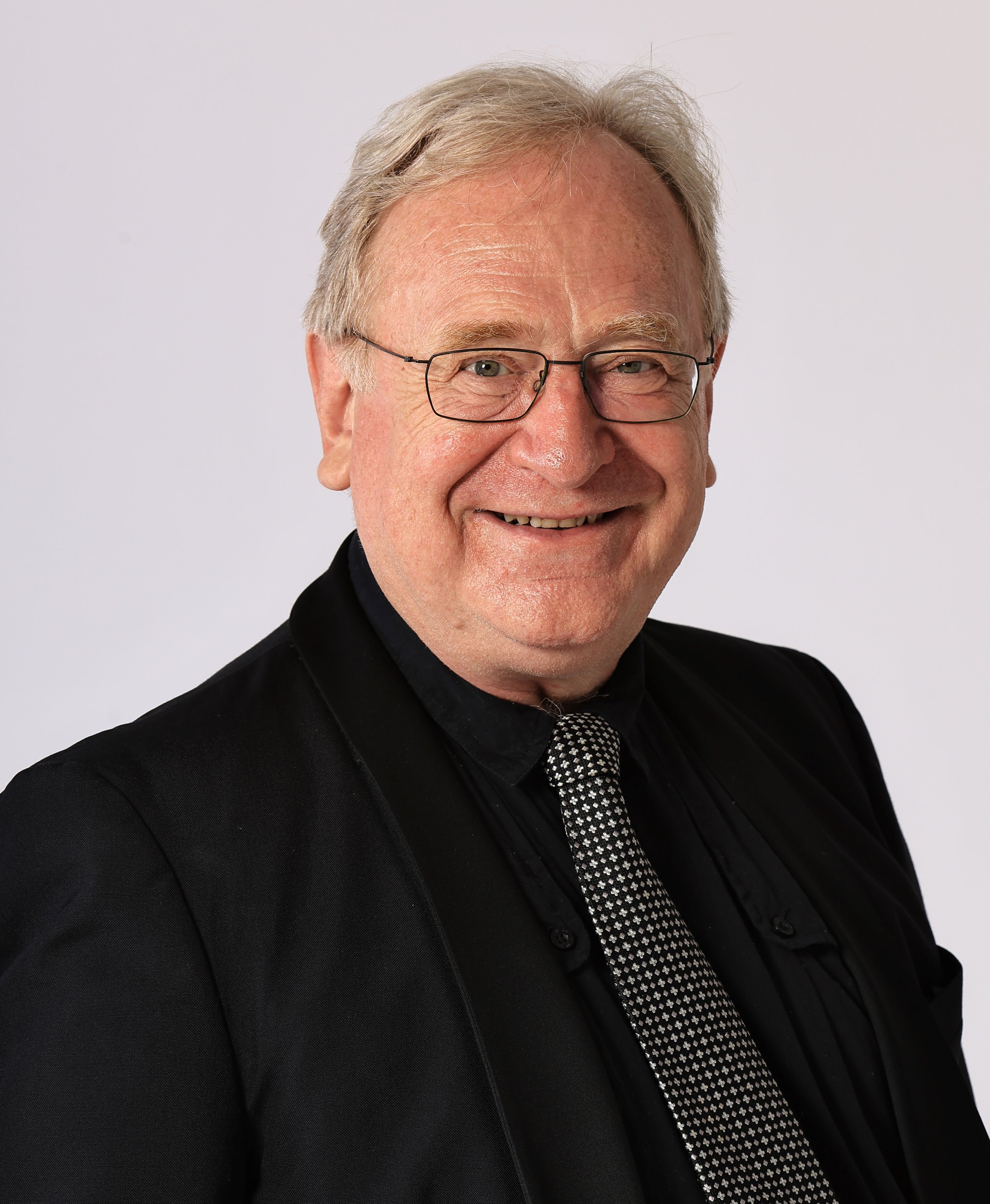 A smiling middle-aged man with glasses and gray hair, wearing a black suit jacket, black shirt, and patterned tie, posing against a plain light background.