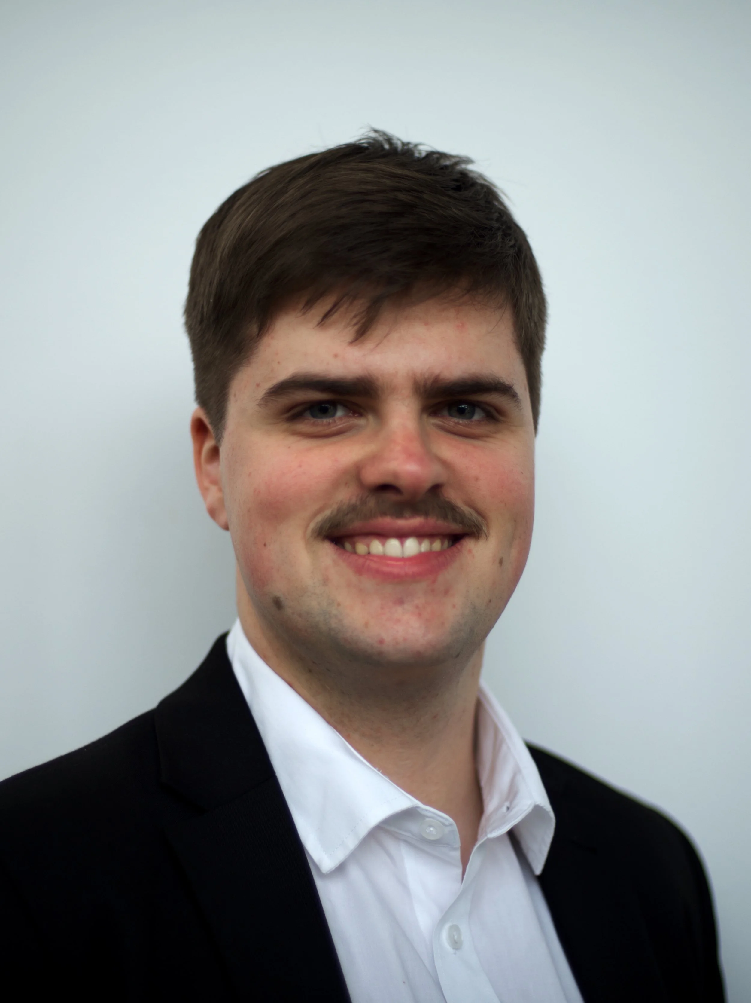 A young man with dark brown hair, blue eyes, and a light complexion, smiling in front of a plain white background. He is wearing a white button-up shirt and a black blazer.