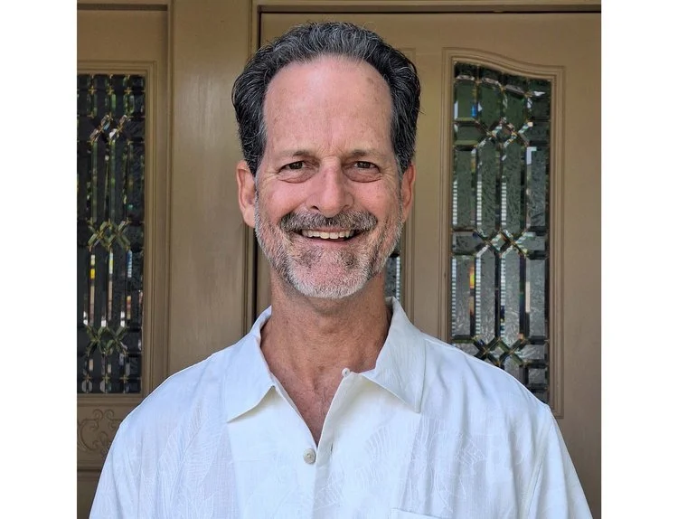 A smiling middle-aged man with gray hair and a beard, wearing a white button-up shirt, standing in front of a door with stained glass windows.
