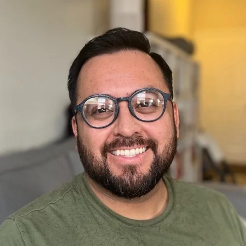 A man with glasses smiling indoors with a blurred background.