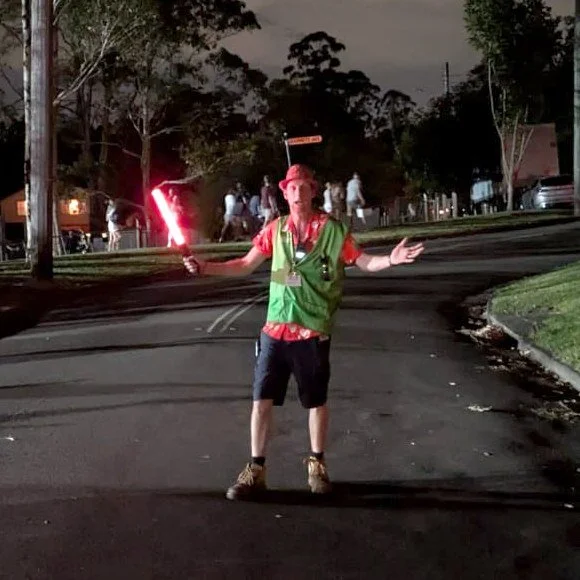 A volunteer in a green vest directing traffic with a light wand on Carols night