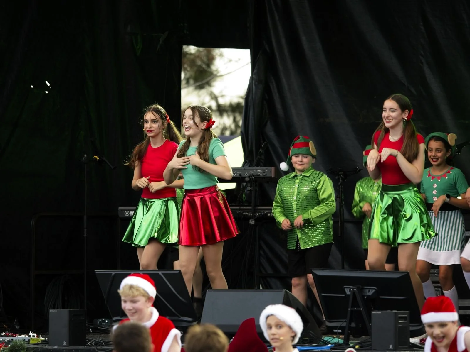 Children performing on stage dressed in holiday-themed costumes, including elves and Christmas attire, with some wearing red and green skirts and others in festive accessories, during a Christmas event.