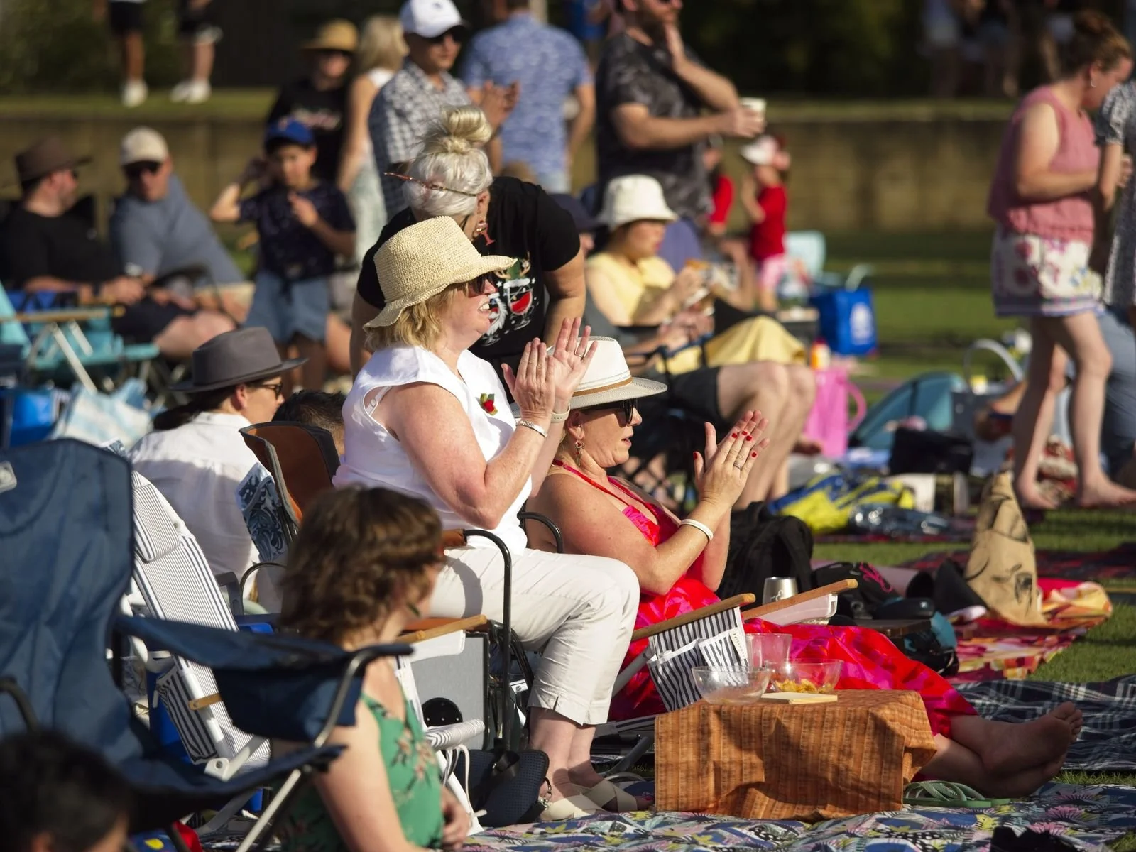 People sitting outdoors on blankets and chairs, many wearing sun hats, watching a performance and clapping.