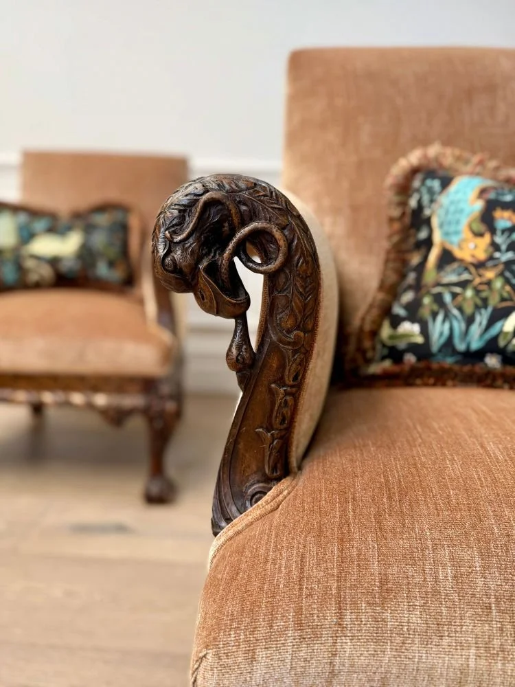 Close-up of an ornate wooden armrest on a tan upholstered sofa with a lever and floral pillow, with a vintage armchair in the background.