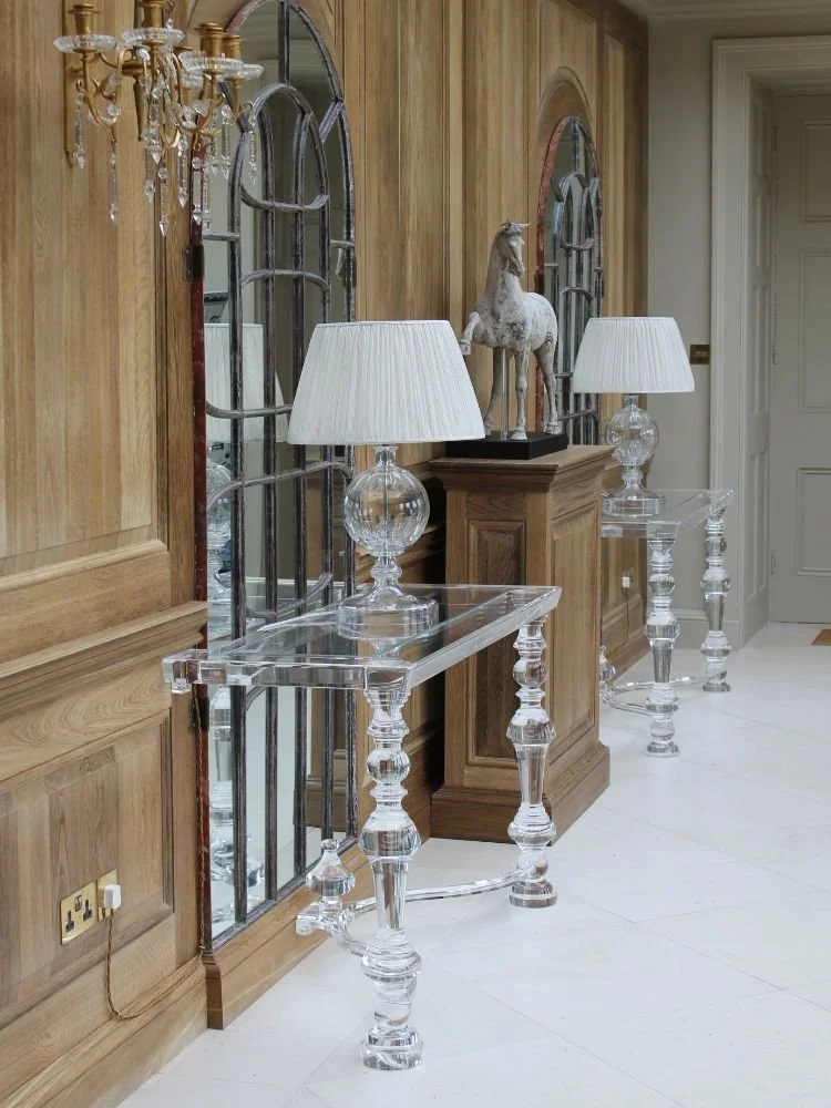 Elegant hallway with wooden wall paneling, three clear glass tables with white lamps, a white horse sculpture atop a wooden cabinet, and large decorative mirrors.