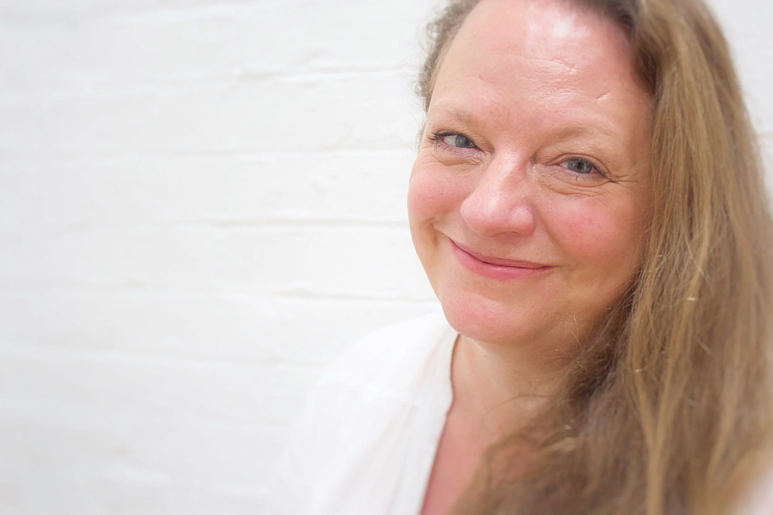 Close-up of a woman with long light brown hair, slightly winking and smiling, against a white background.