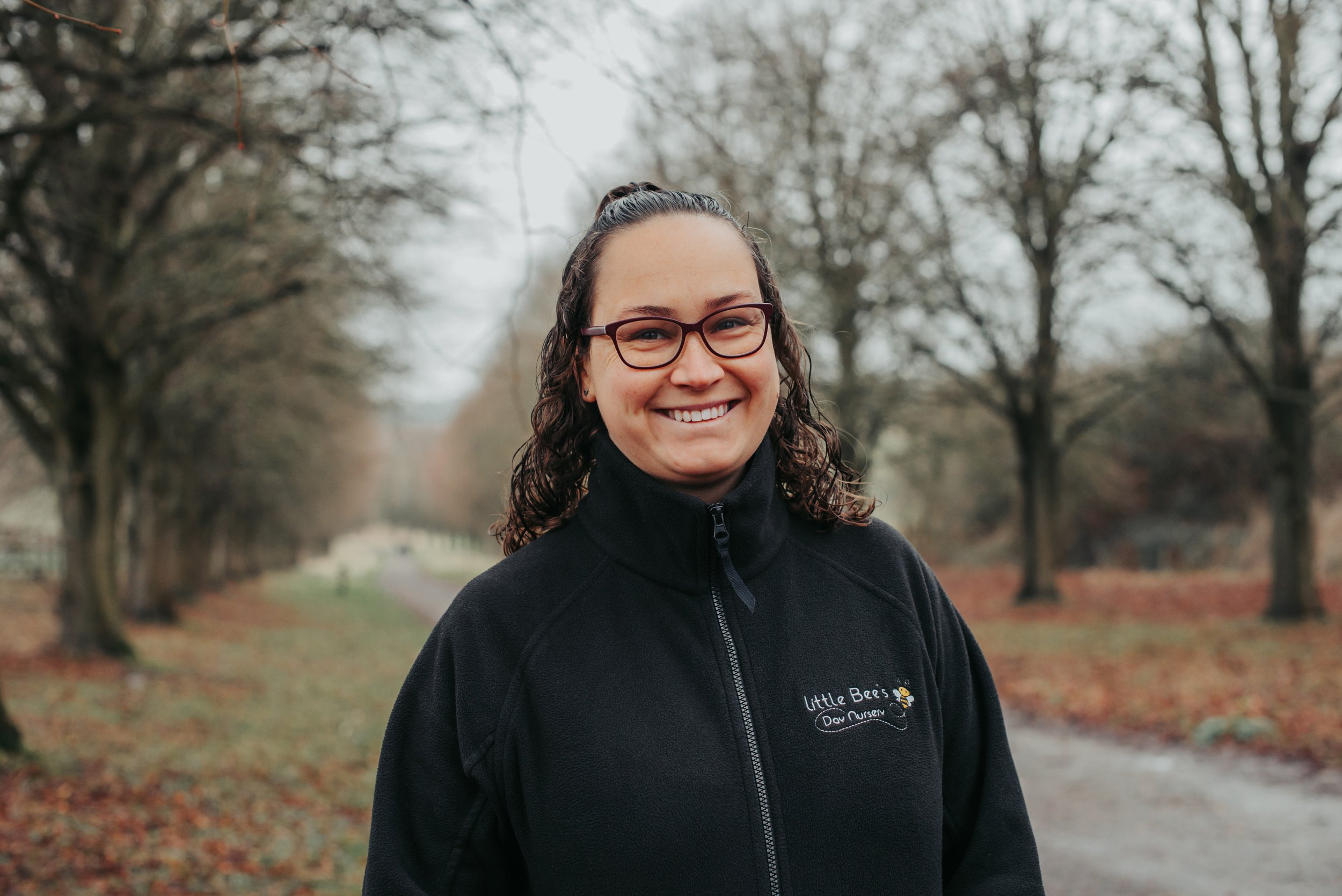 A woman with brown and reddish hair, blue eyes, and multiple facial piercings smiling outdoors in front of green foliage. She is wearing a black fleece jacket with a logo that reads 'Little Bee's Day Nursery' and has a small bee illustration.