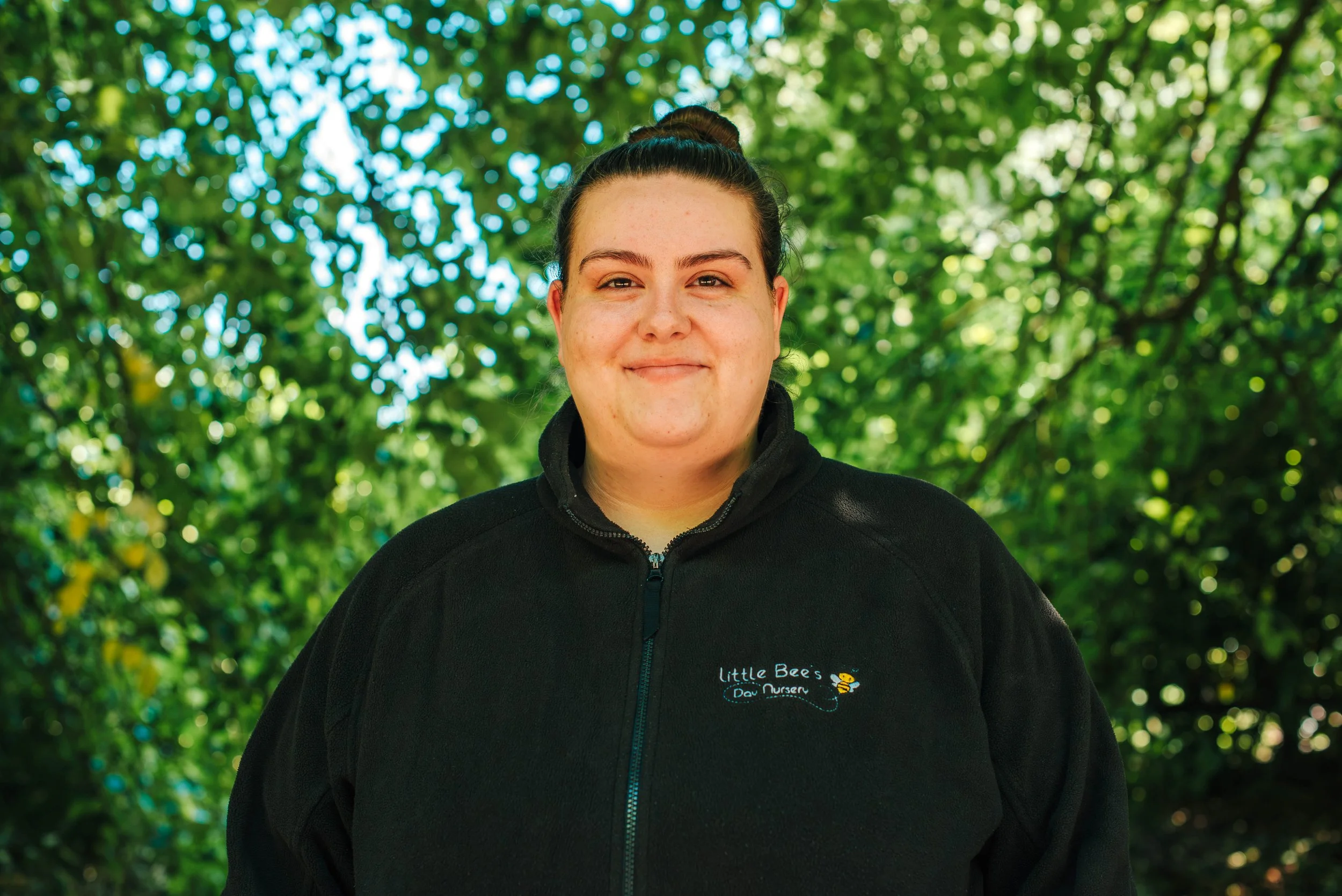A young woman smiling outdoors in front of green foliage, wearing a light gray polo shirt with the logo 'Little Bee's Dan Nursery' and a bee graphic.