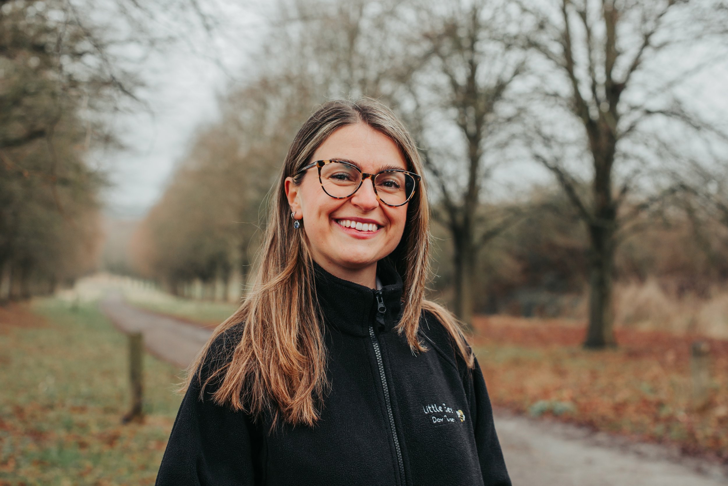 A smiling woman with glasses and earrings standing outdoors in front of green foliage, wearing a black fleece jacket with an embroidered logo that reads 'Little Bee's' and a graphic of a bee.