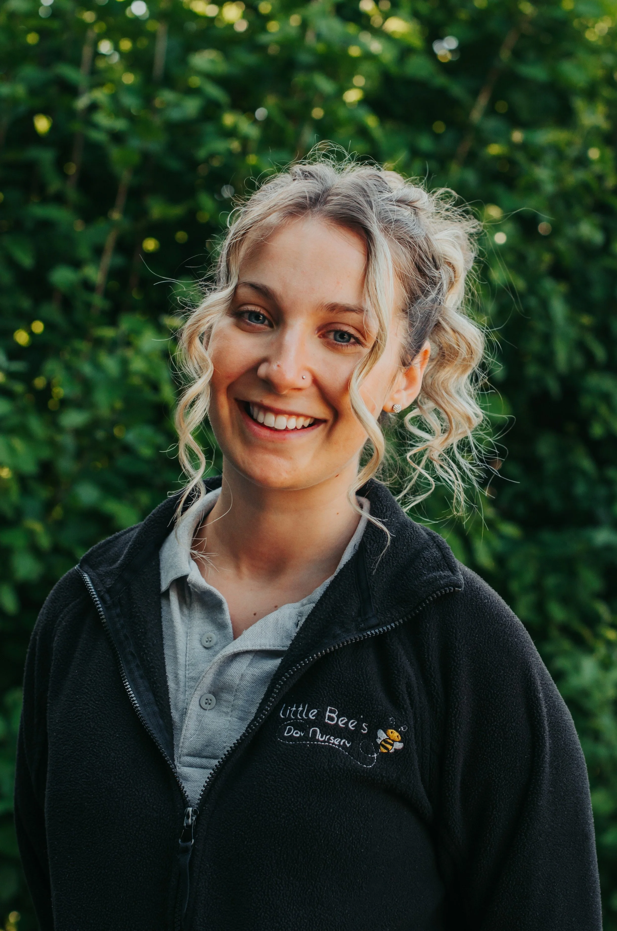 A young woman with long blonde hair, smiling, wearing a gray polo shirt with a bee logo and the words "Bees" on it, standing in front of a white wall.