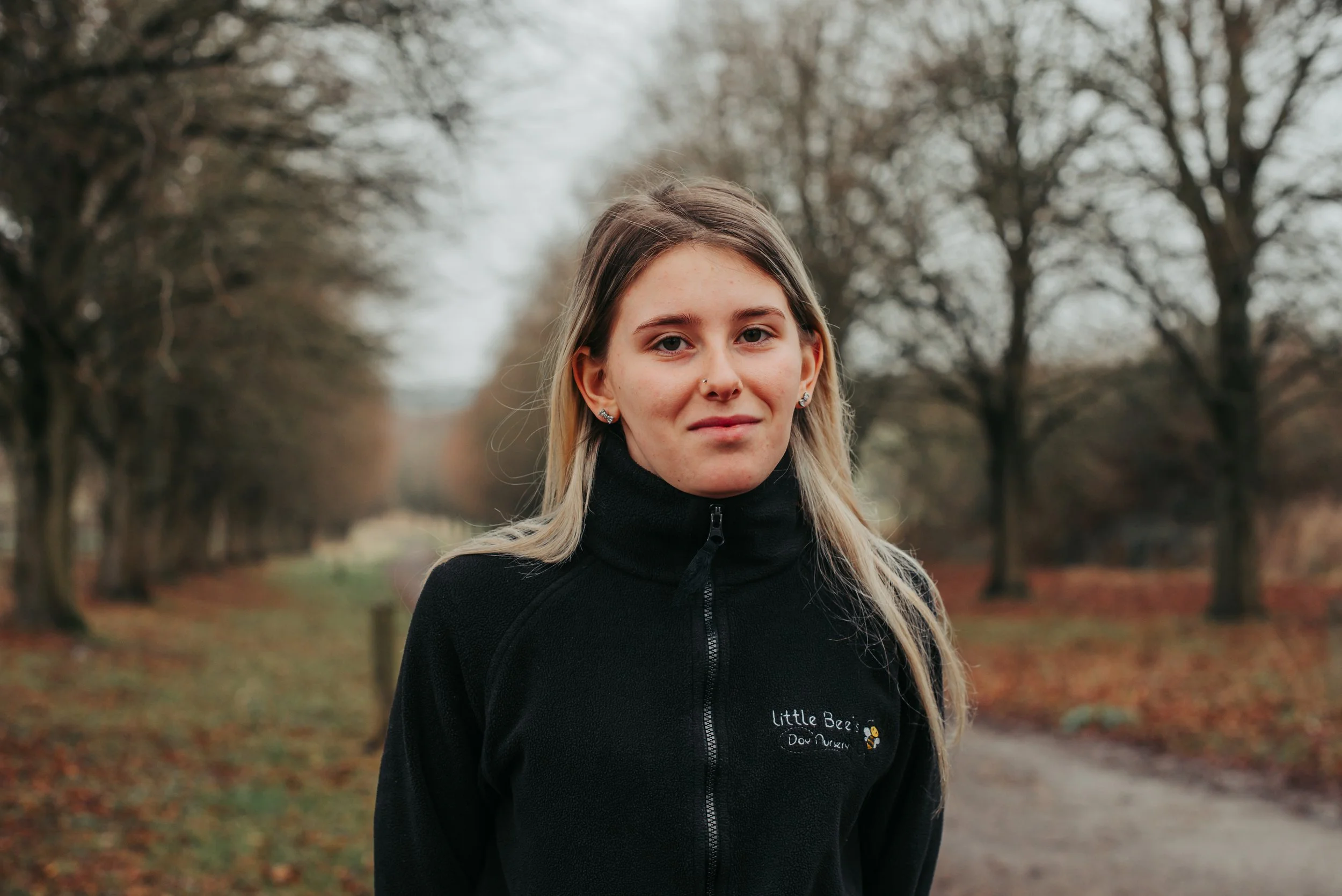 Young woman wearing a light gray polo shirt with 'Little Bee's Day Nursery' logo, standing against a white wall.