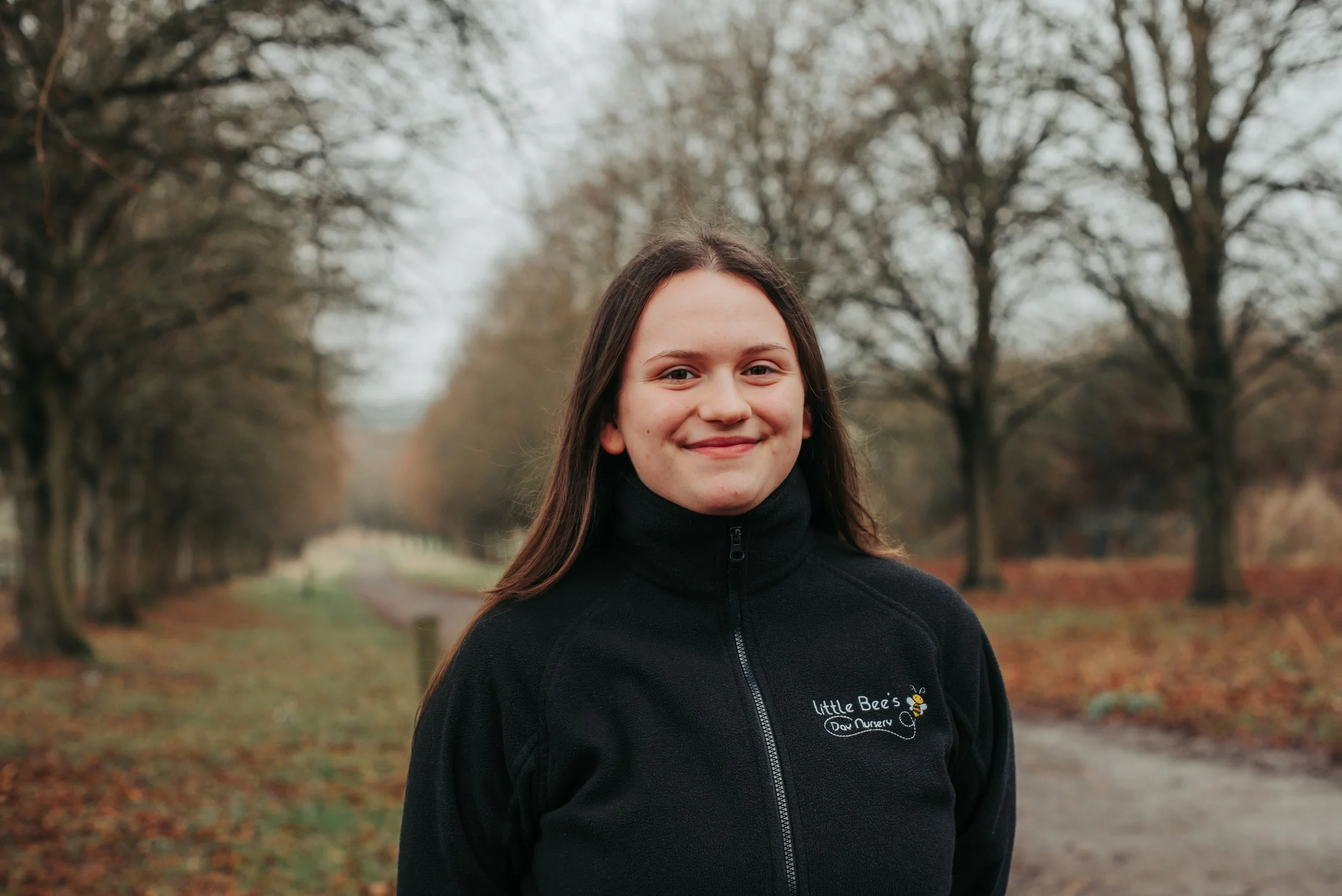 A young woman with long dark hair, glasses, and a smile standing outdoors in front of green foliage.