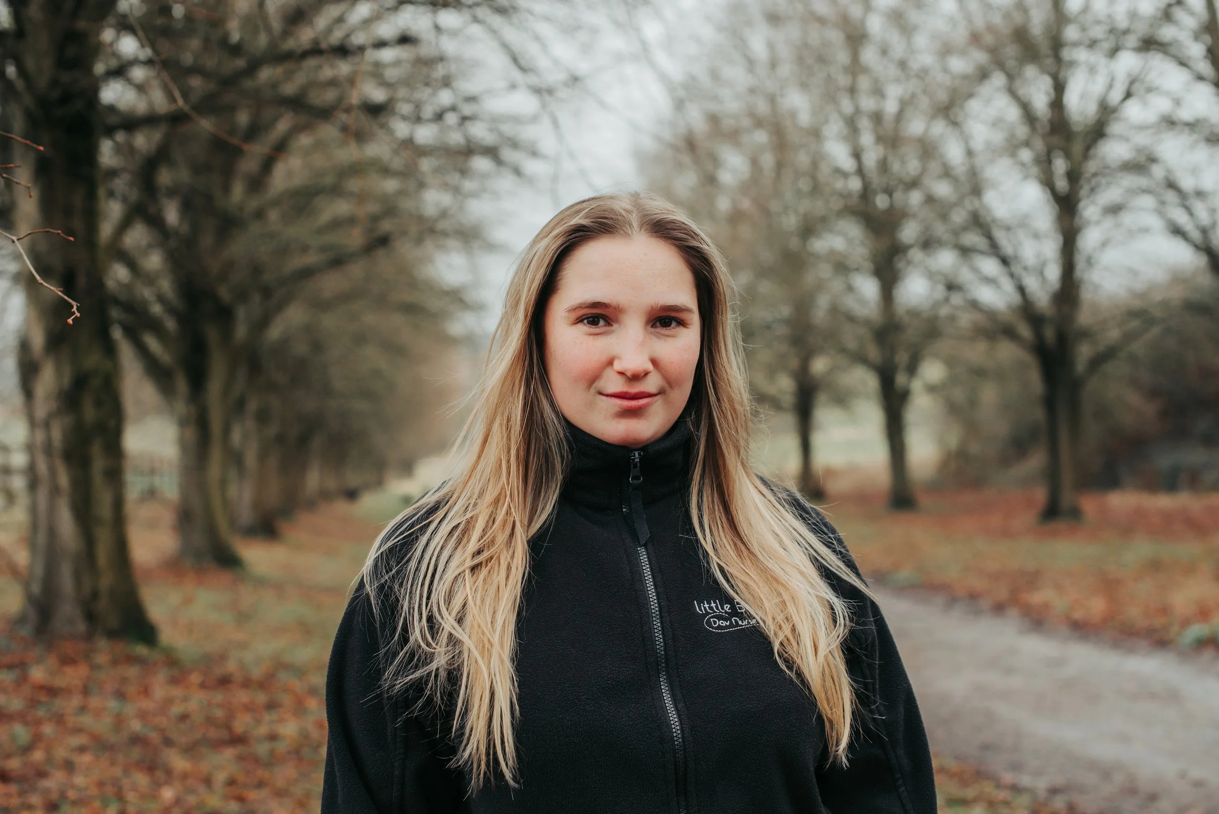 A young woman with light brown hair in a braid, wearing a black zip-up jacket with a embroidery of a bee and the words 'Little Bee's Day Nursery,' smiling outdoors with trees in the background.