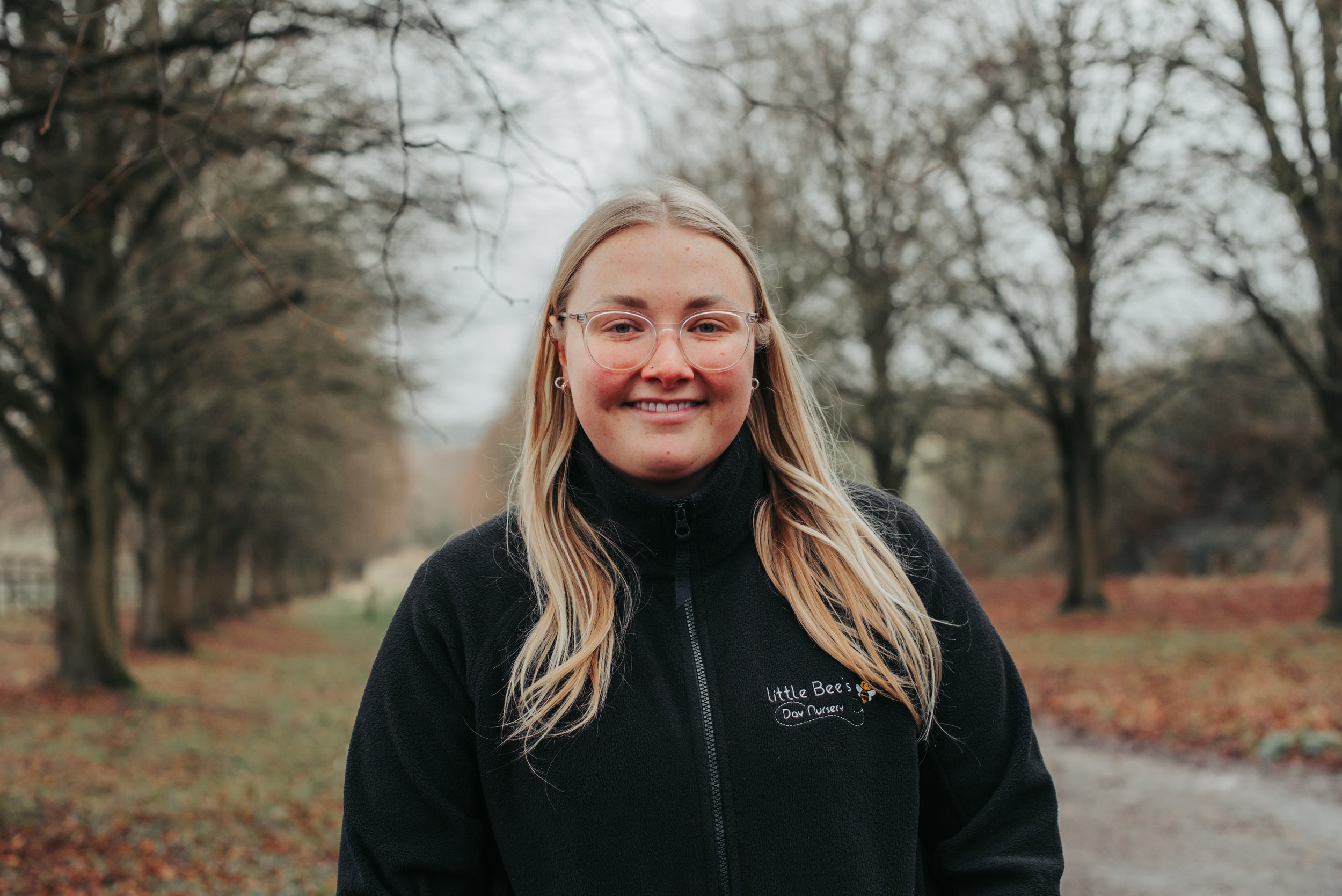 A young woman with long blonde hair and a light complexion, wearing a black zip-up jacket and a gray shirt, standing in front of a plain white wall.