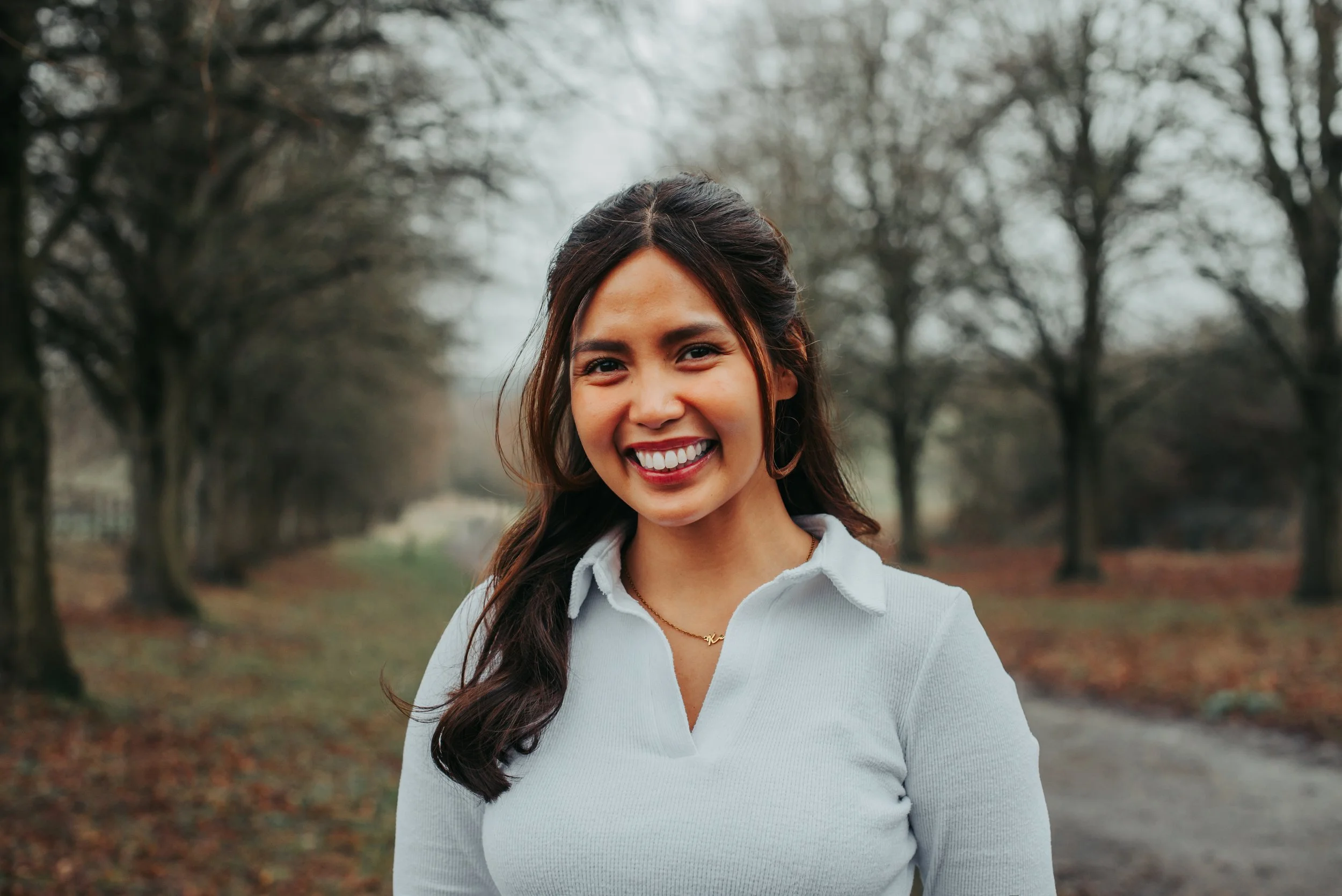 A woman smiling outdoors, wearing a black button-up sweater and a necklace that says 'Kealoha', with dark hair and a background of green foliage.