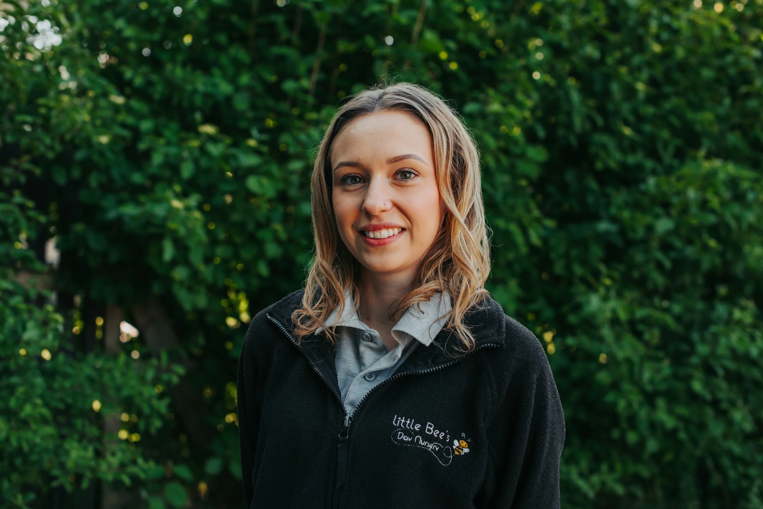 Young woman with blonde hair, glasses, and a black jacket standing against a plain white wall.