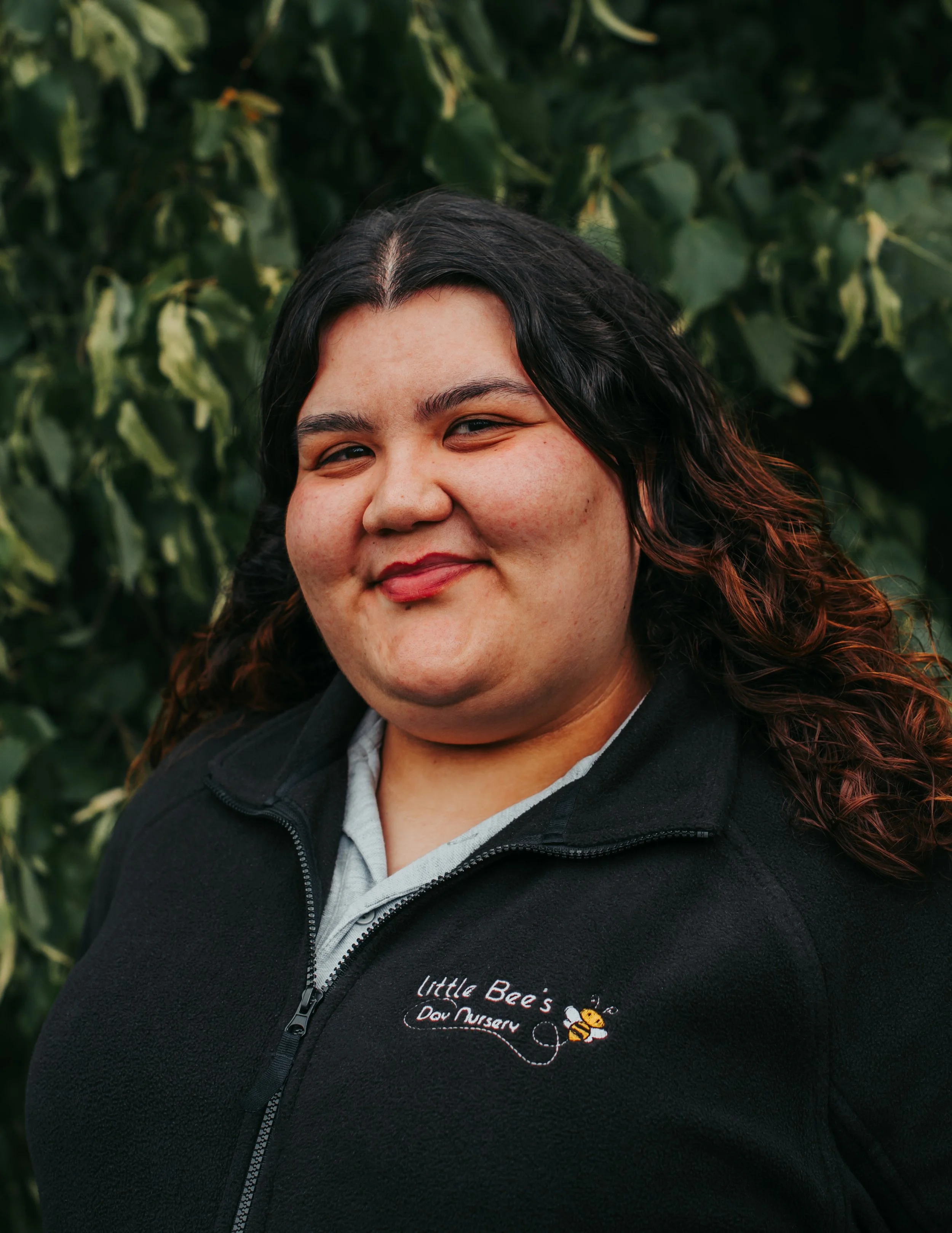 Woman with glasses smiling outdoors on a path with trees in the background, wearing a black fleece jacket with a logo that reads 'Little Bees Day Nursery'.