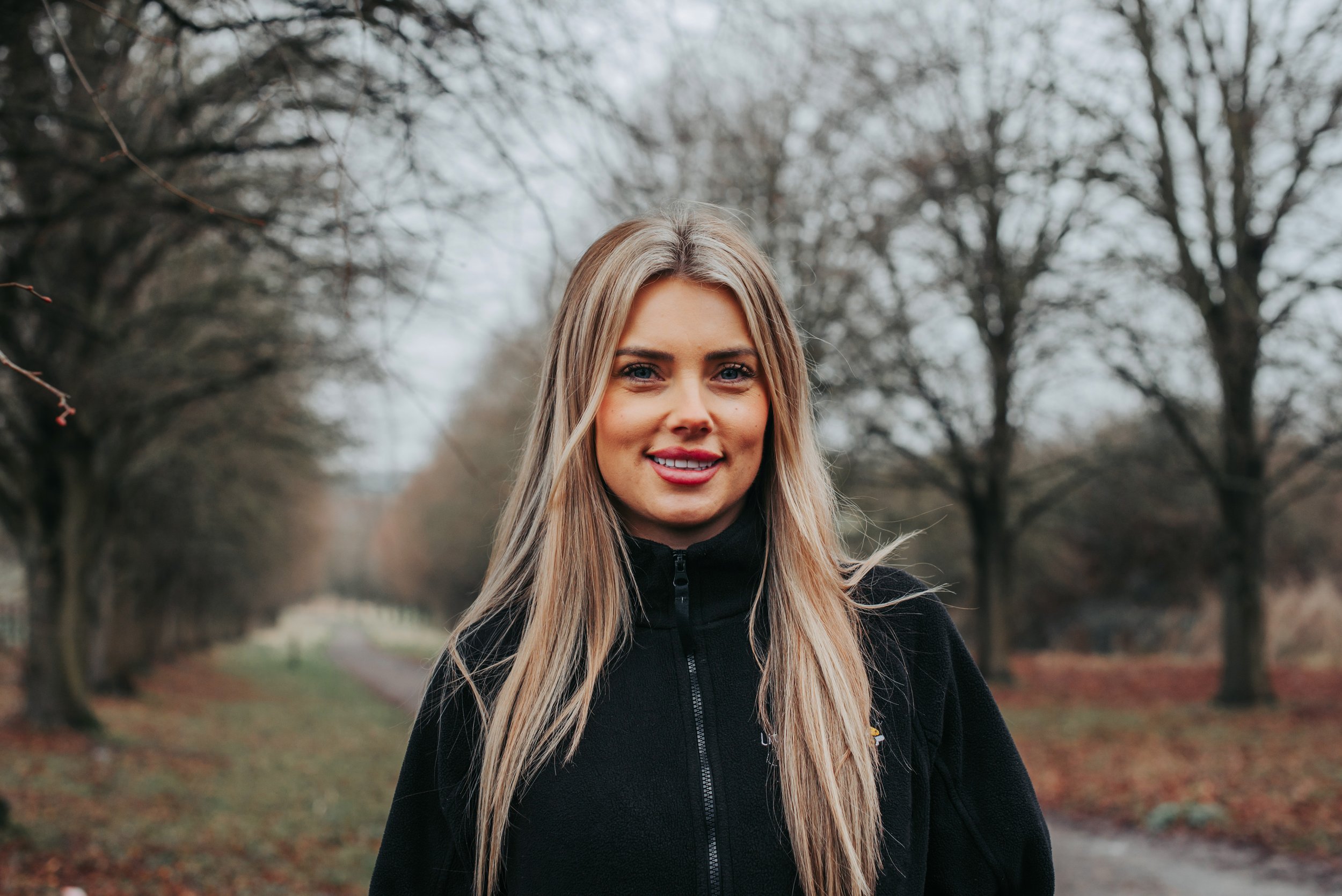 A smiling woman with blonde hair and blue eyes standing outdoors in front of green foliage, wearing a black jacket with a logo that says 'little Bee's Doo Nursery' and a gray collared shirt.