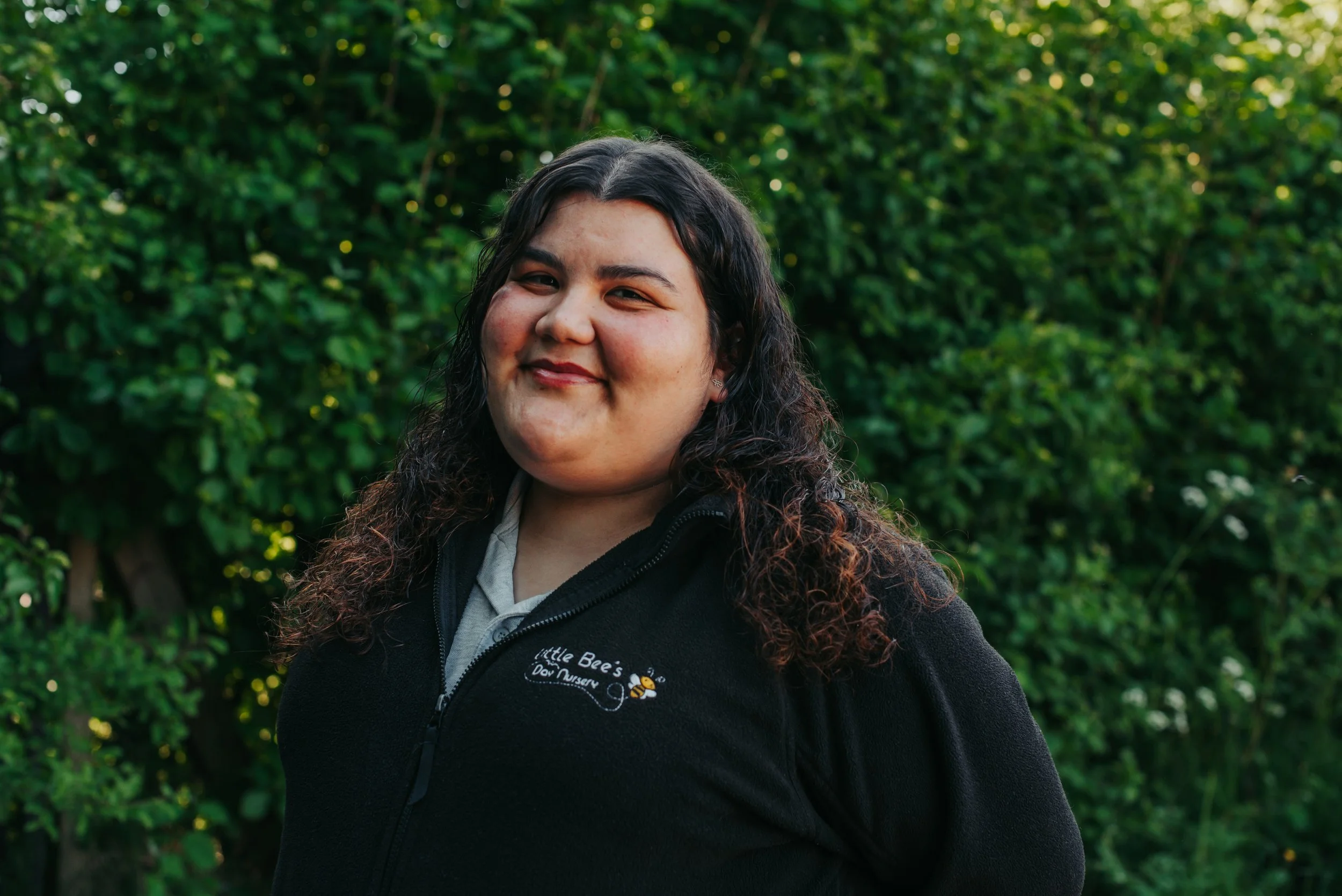 A woman with dark wavy hair smiling outdoors, wearing a black jacket with the logo 'Little Bee's Day Nursery' and a bee graphic, against a background of green foliage.
