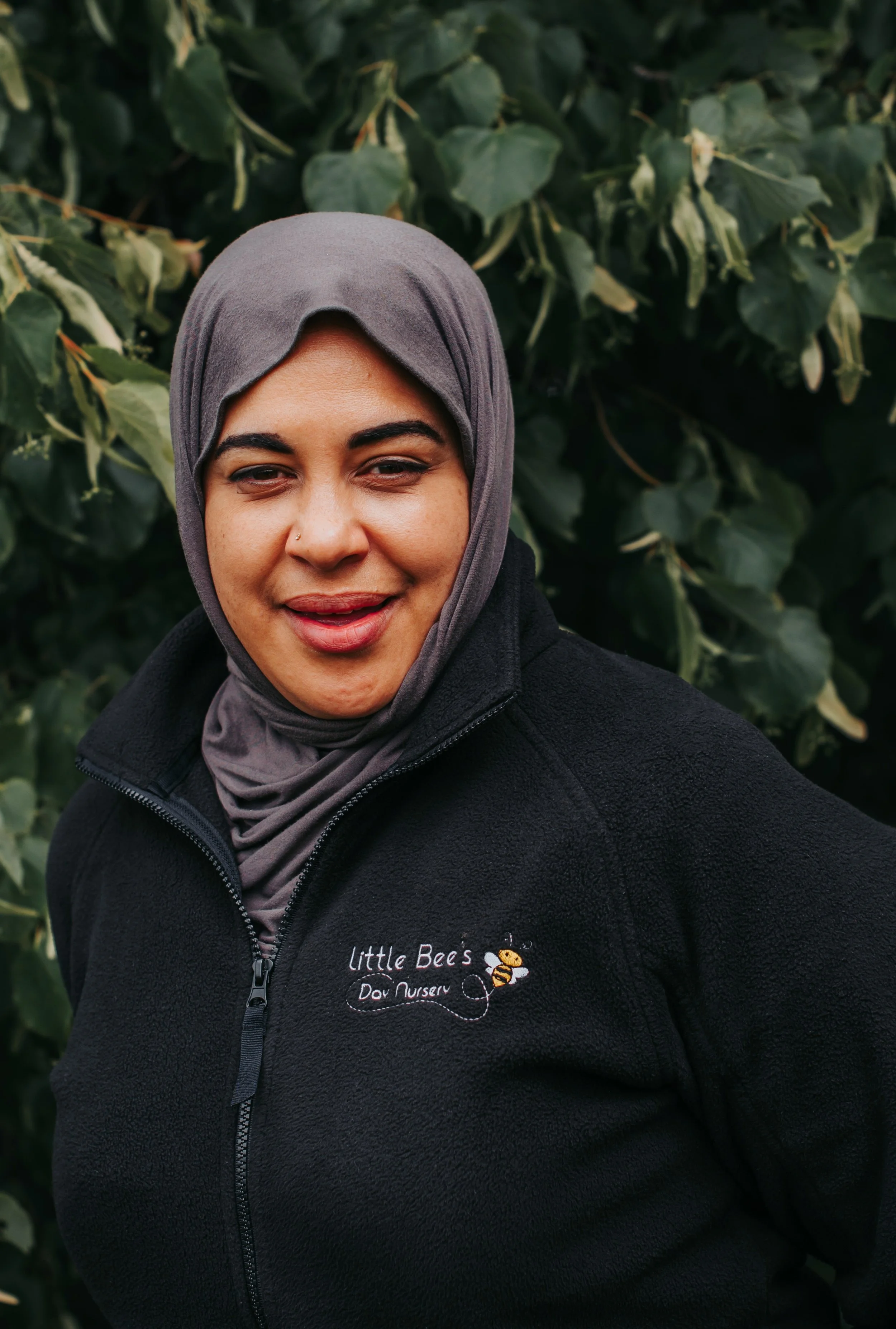 A woman with long, blonde hair wearing a black jacket and a gray shirt, standing outdoors in front of green leafy plants.