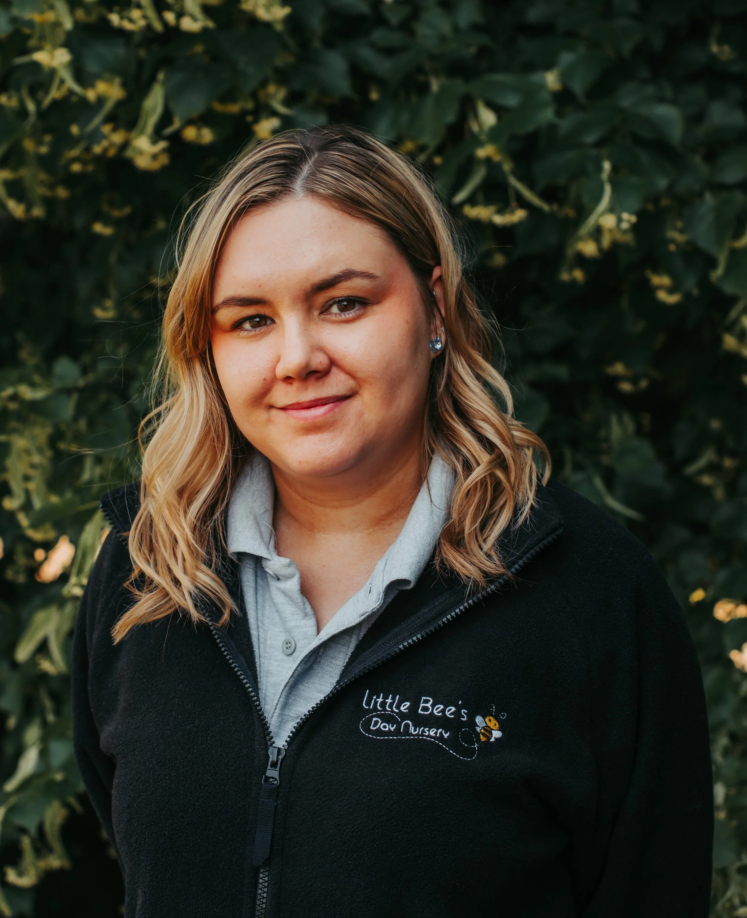 Woman with glasses smiling outdoors on a path with trees in the background, wearing a black fleece jacket with a logo that reads 'Little Bees Day Nursery'.