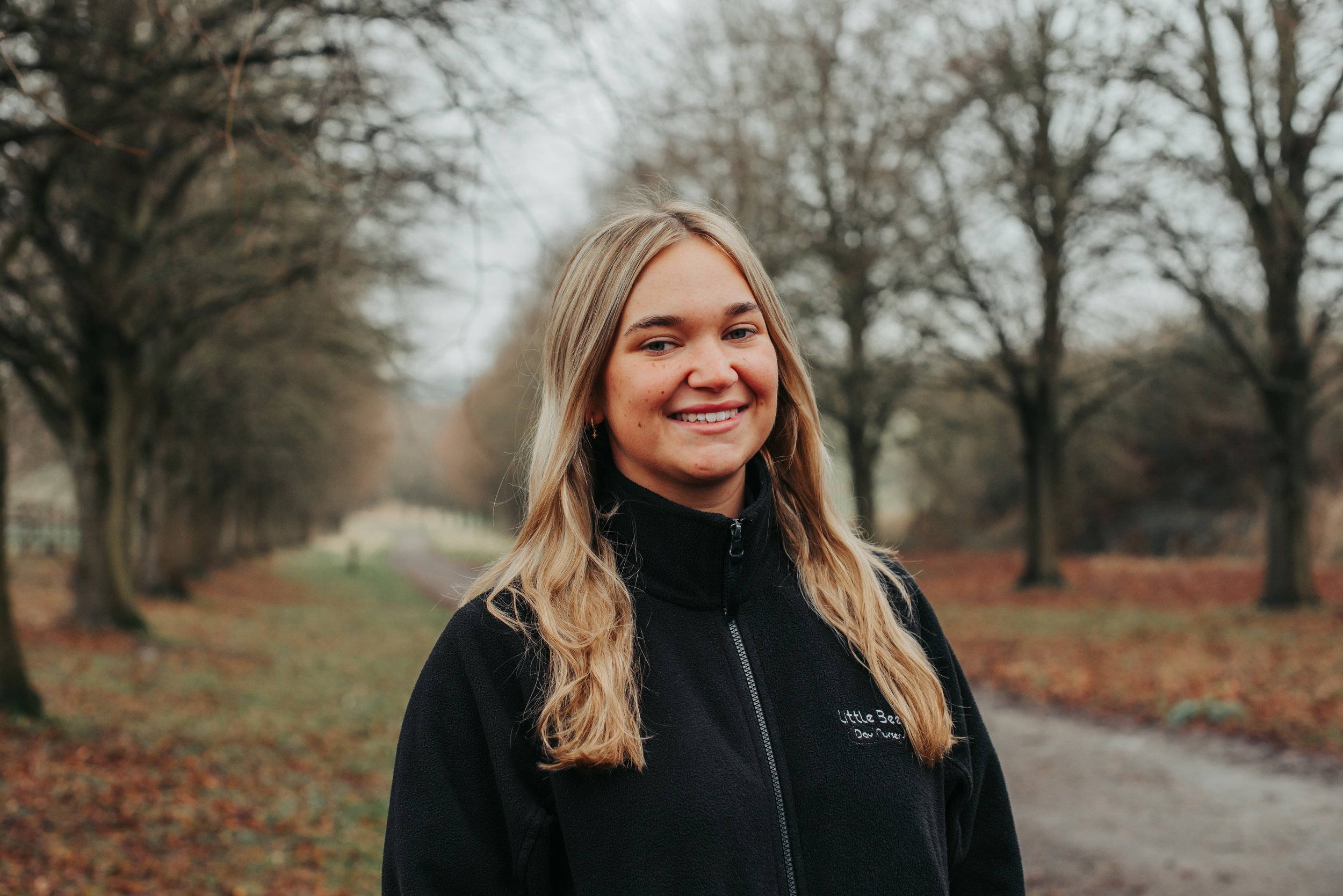 A young woman with long blonde hair, blue eyes, and light skin standing outdoors with a blurred green background. She is wearing a black zip-up fleece jacket with embroidered text that reads 'Bees Little Bees Do Nursery' and a small bee logo.