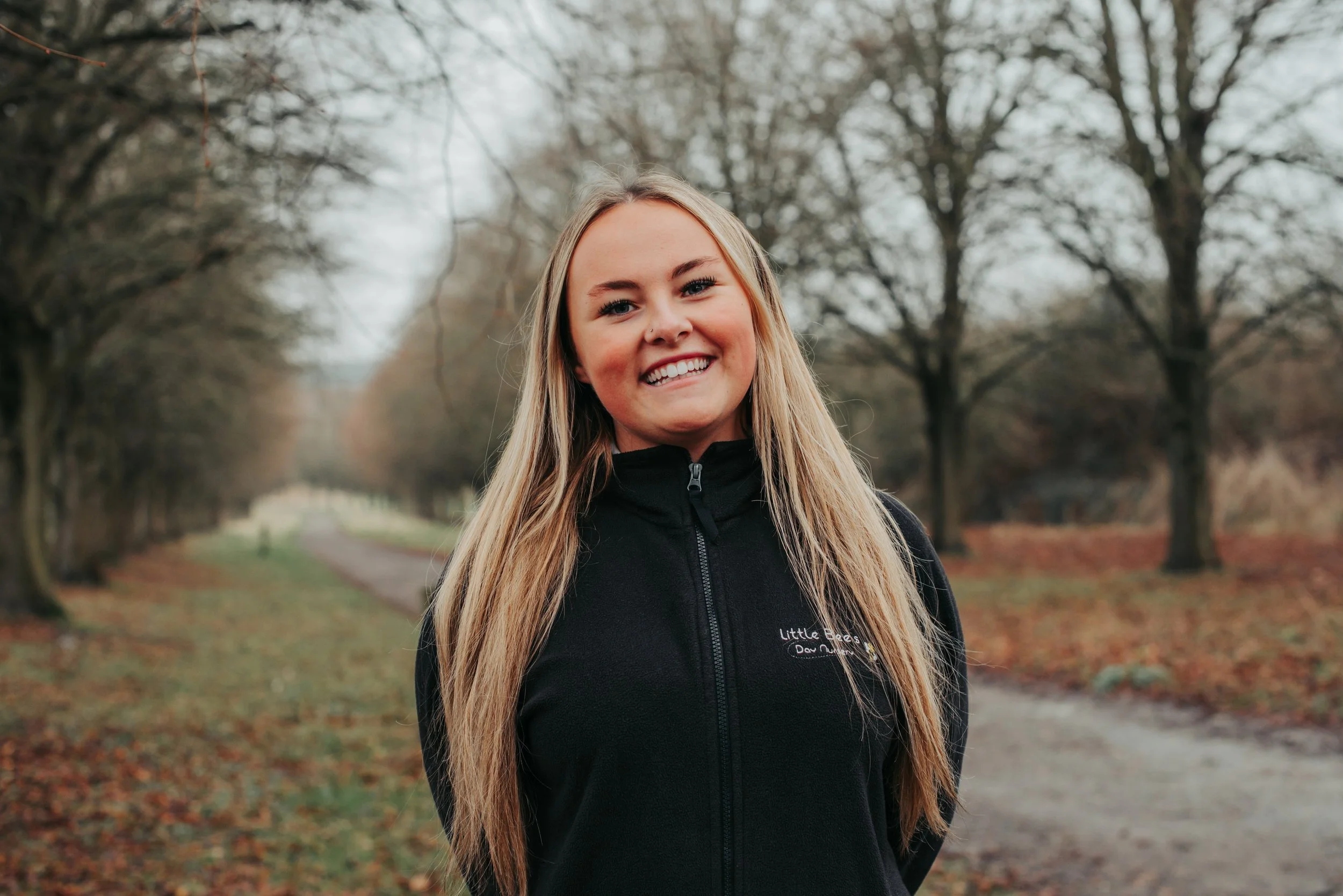 A smiling woman with blond hair, wearing a black jacket with 'Little Bee's Day Nursery' embroidered on it, outdoors with blurred trees in the background.