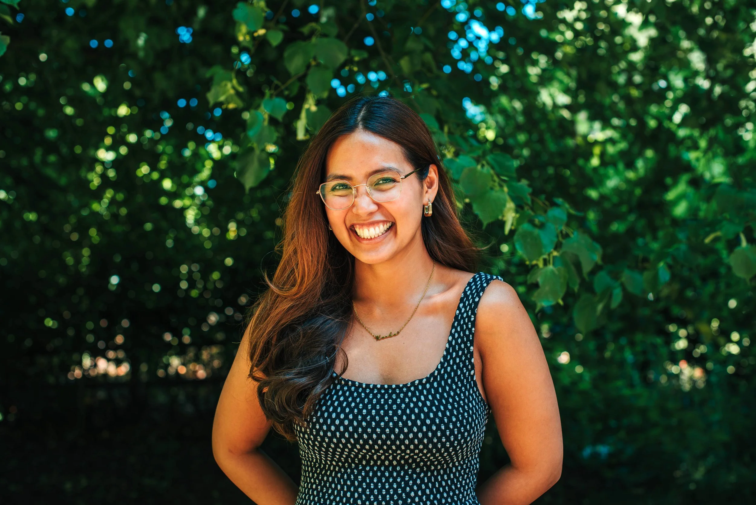 A woman smiling outdoors, wearing a black button-up sweater and a necklace that says 'Kealoha', with dark hair and a background of green foliage.