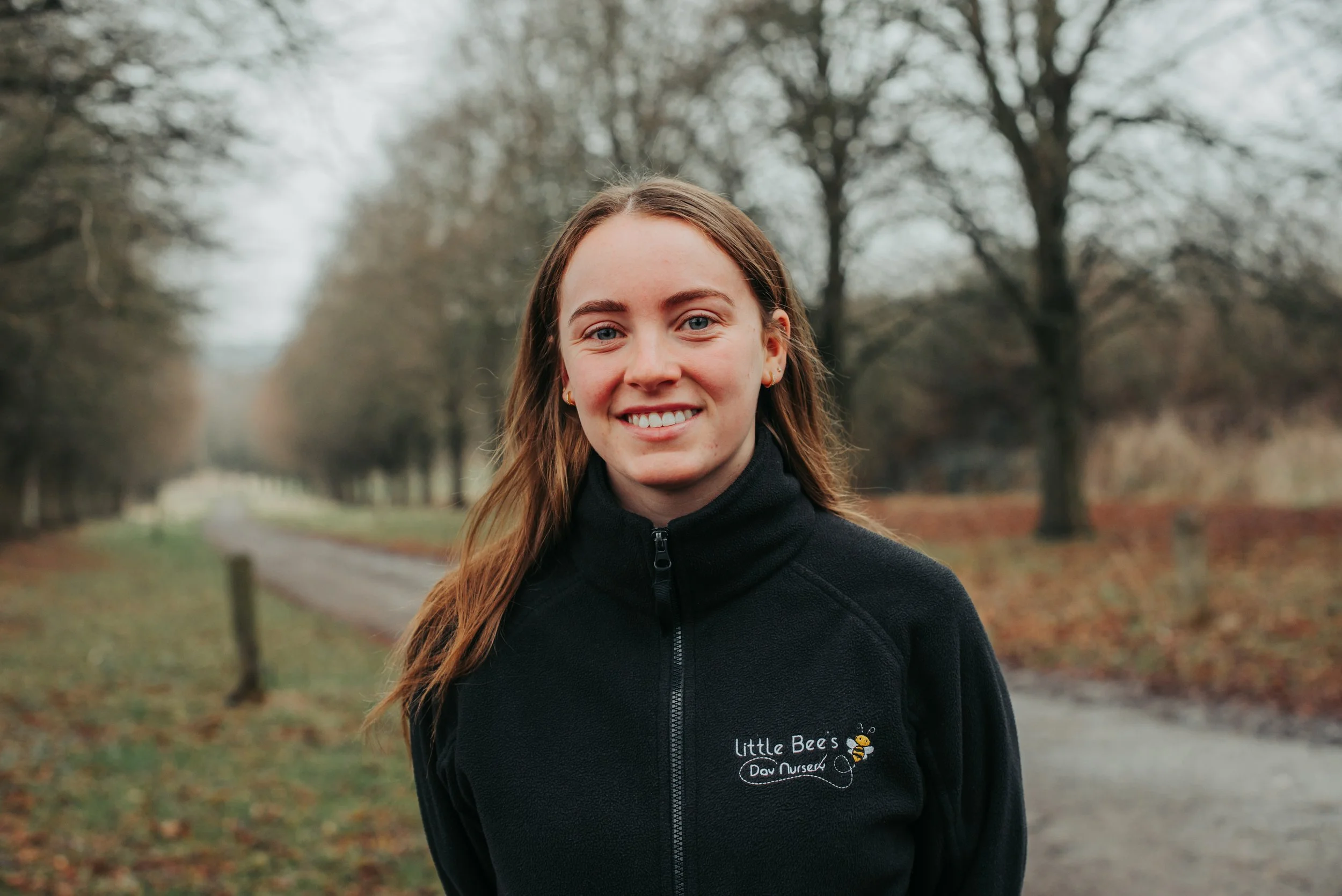 A young woman with light brown hair and fair skin, wearing a light blue polo shirt, standing outdoors in front of lush green foliage and red flowers.