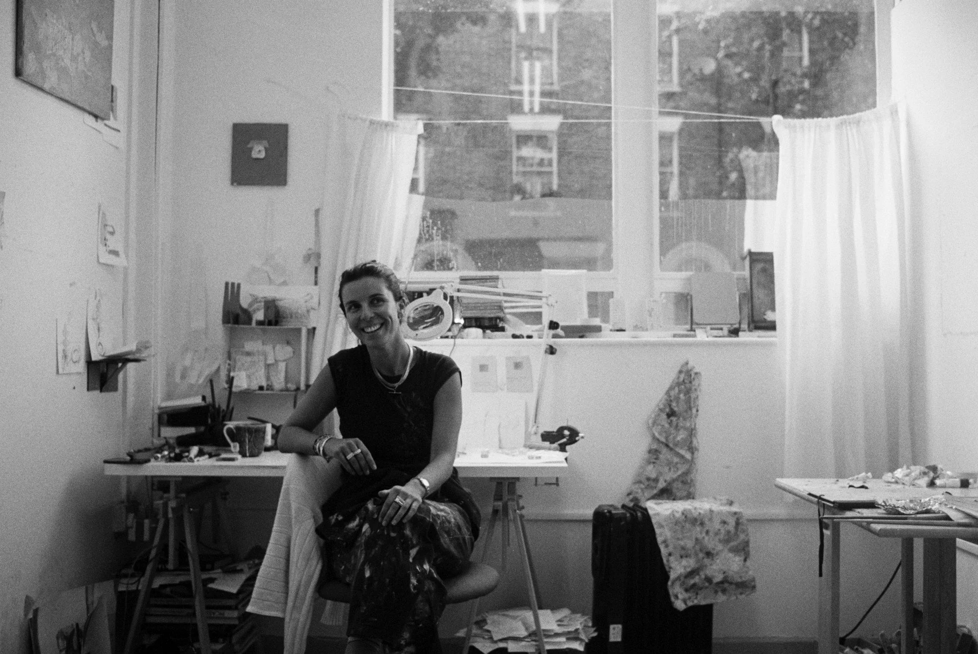 A woman sitting on a chair in a cluttered art studio or workspace, smiling at the camera, with various art supplies and papers around her.