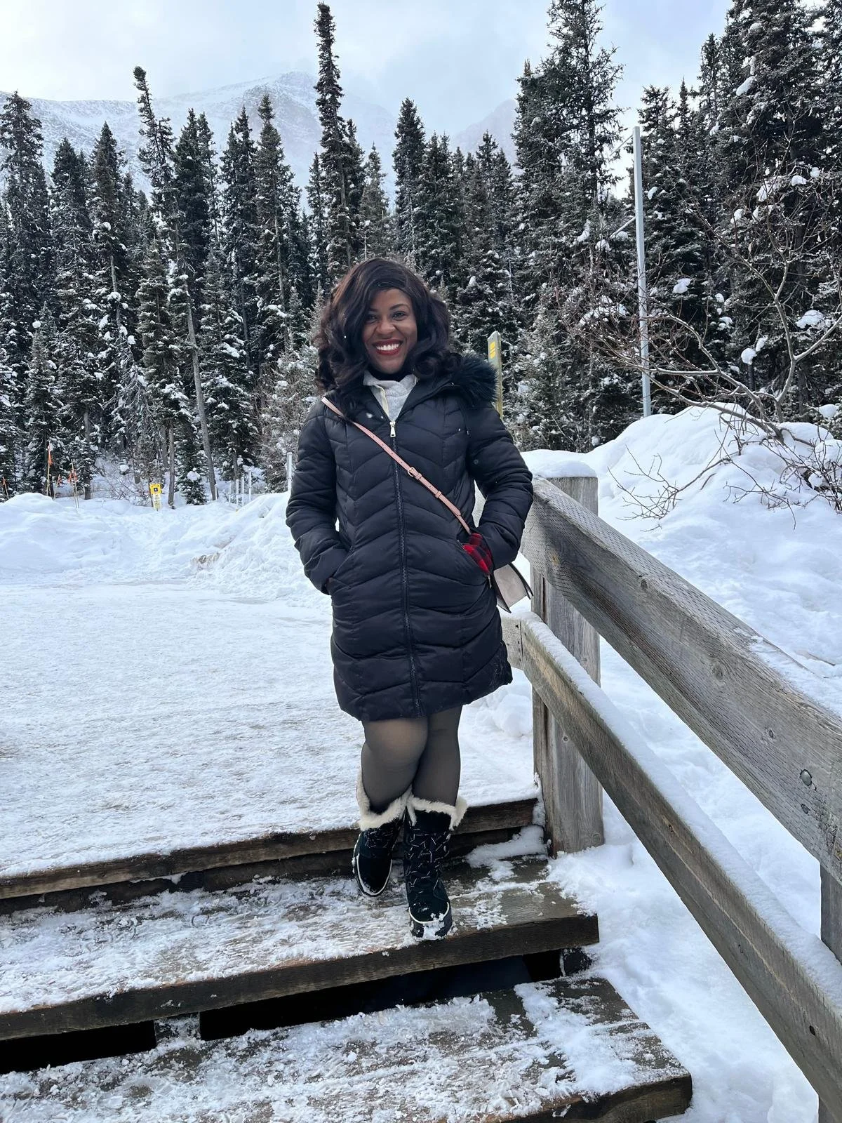 A woman standing on a snow-covered wooden platform in a snowy forest with tall pine trees and mountains in the background, smiling at the camera, wearing a black winter coat, gray tights, and snow boots.