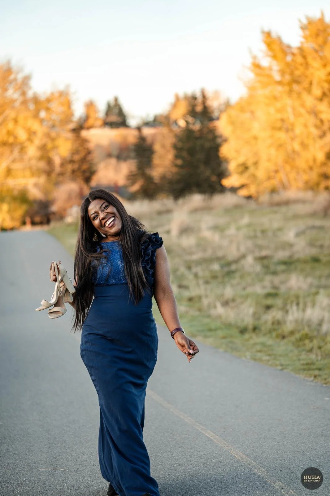 A woman in a blue dress walking on a pathway, holding high heels, smiling and laughing with trees and a sunset in the background.