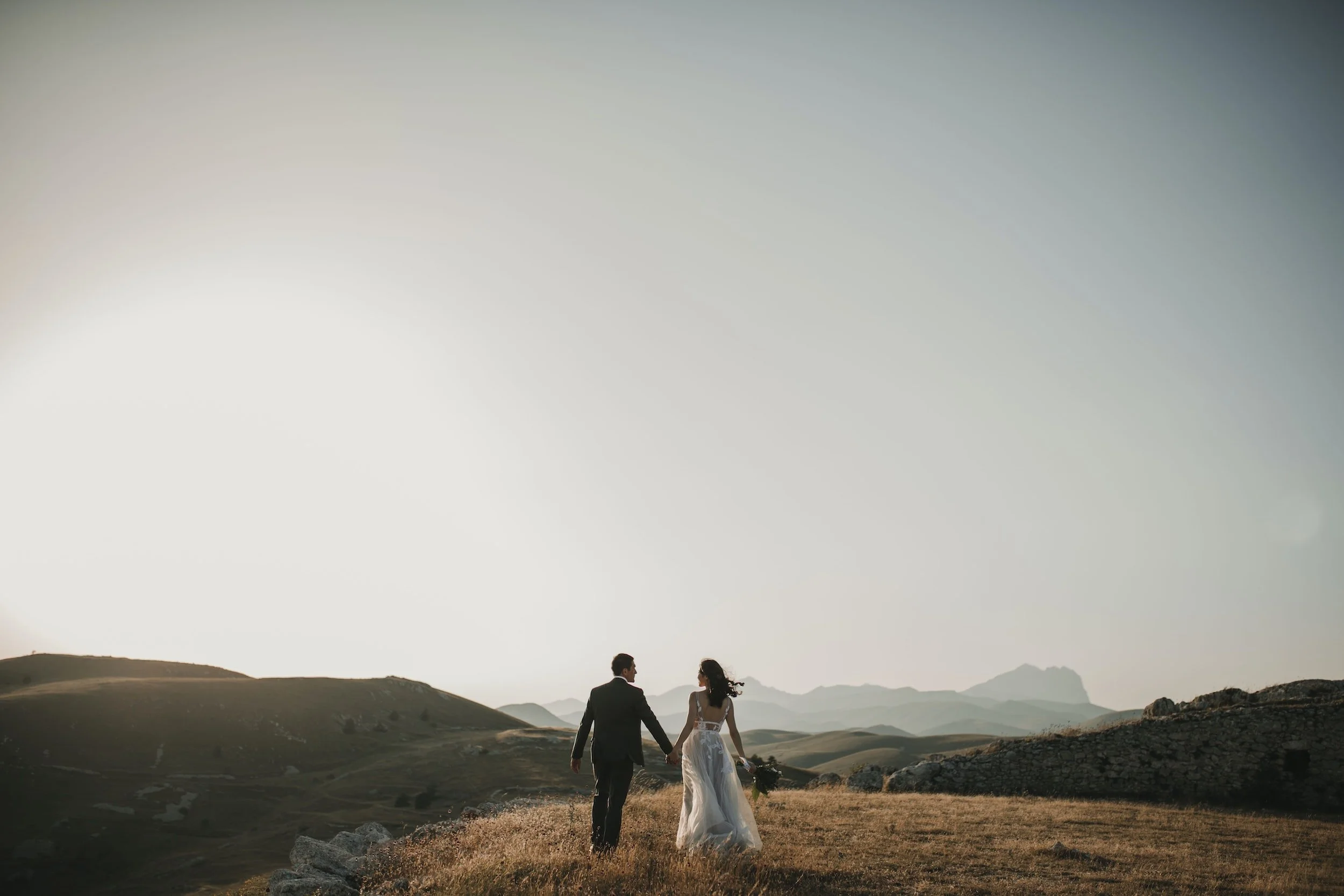 A bride and groom walk hand in hand in a scenic outdoor landscape during sunset.