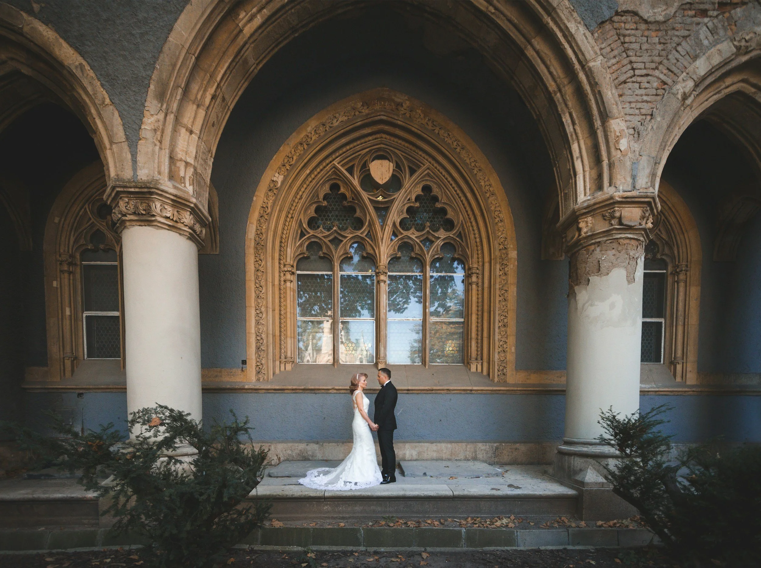 A bride and groom in wedding attire standing face to face in front of a large gothic-style arched window with intricate stonework, inside a historic building with columns and dark walls.