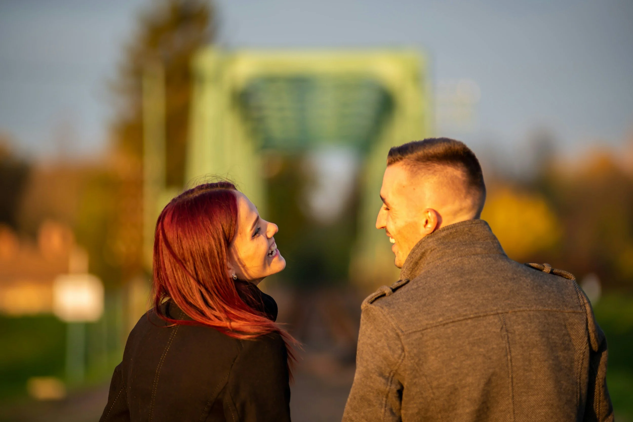 A woman with red hair and a man in military uniform smiling and facing each other outdoors at sunset.