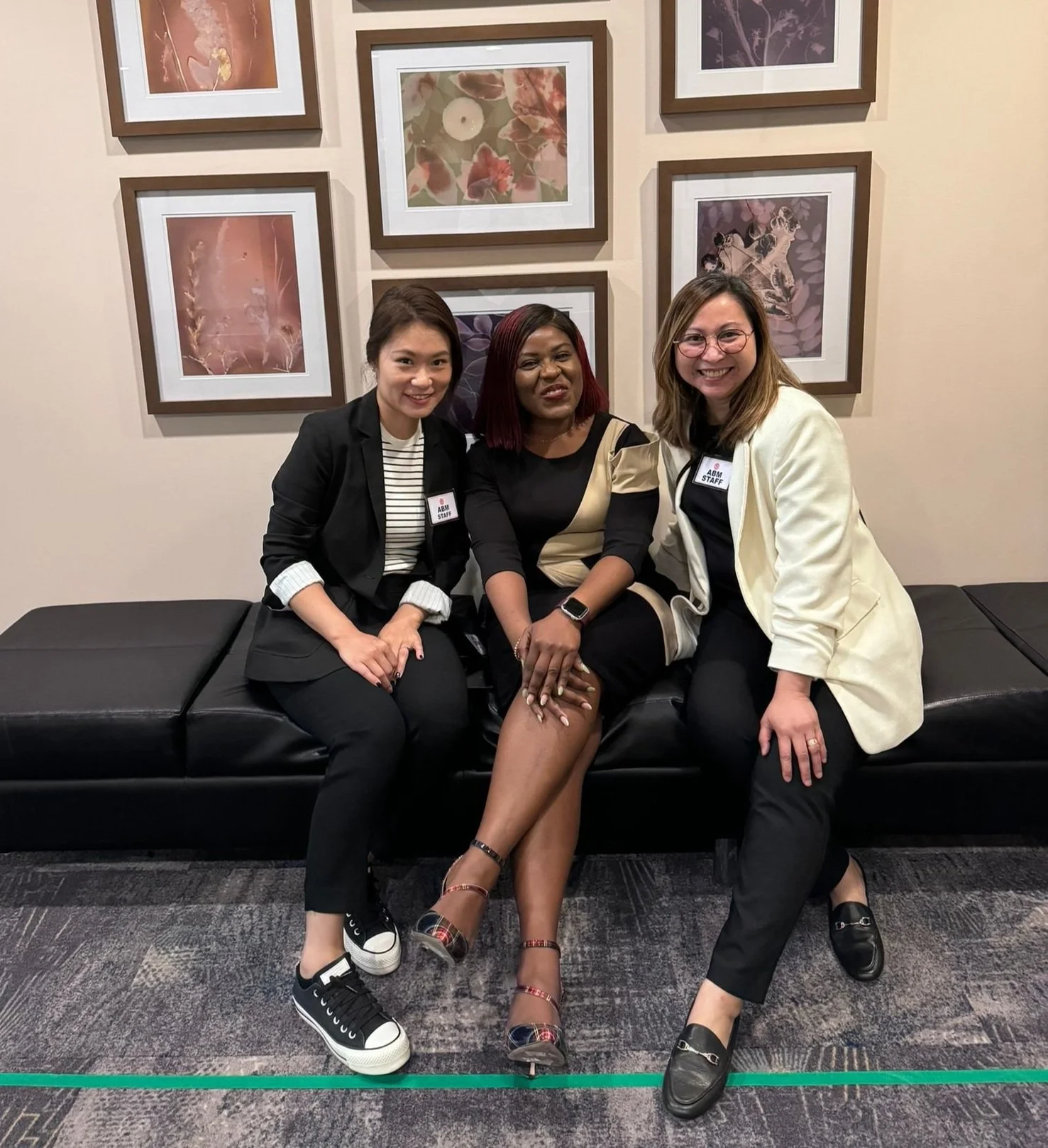Three women sitting on a black bench in front of a beige wall with framed artwork. The woman in the middle has dark skin and red hair, wearing a black and beige dress, with her hands resting on her lap. The woman on the left has light skin and dark hair, dressed in black with a striped top, and is smiling at the camera. The woman on the right has light skin and light brown hair, wearing glasses, a white blazer, and black pants, also smiling. All three are wearing black shoes, and the two women on the ends have 'Staff' name tags.