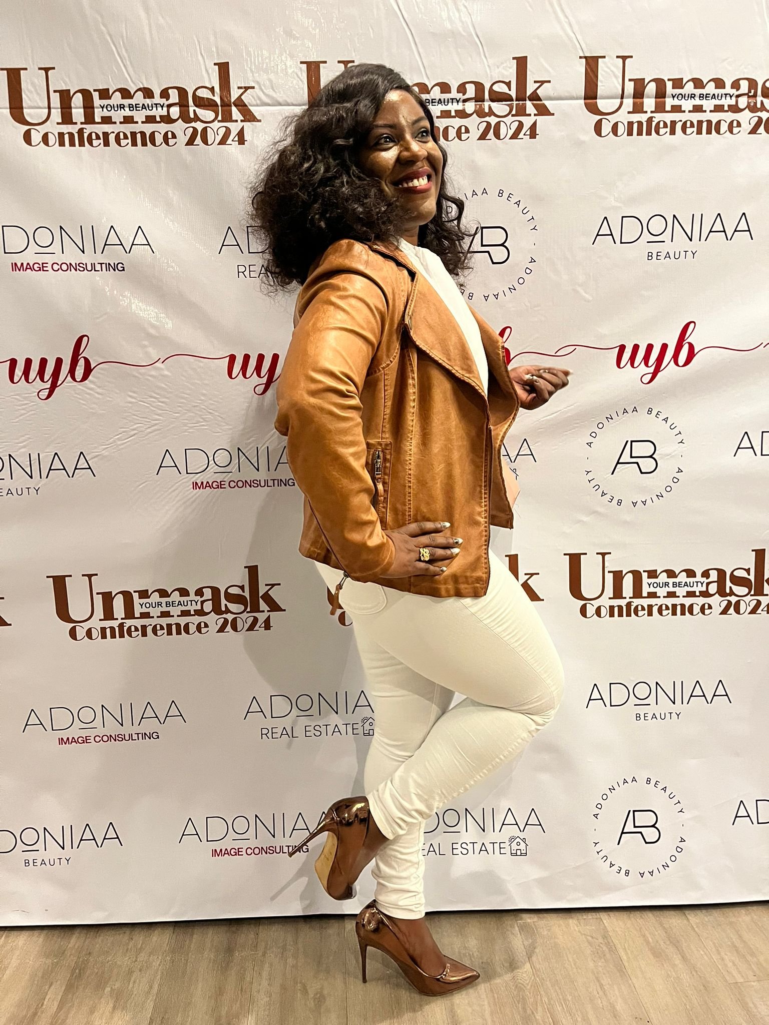 A woman posing confidently in front of a backdrop at the Unmask Conference 2024. She is wearing a tan leather jacket, white pants, and brown high-heeled shoes, smiling and standing with one foot on the ground and the other knee bent.