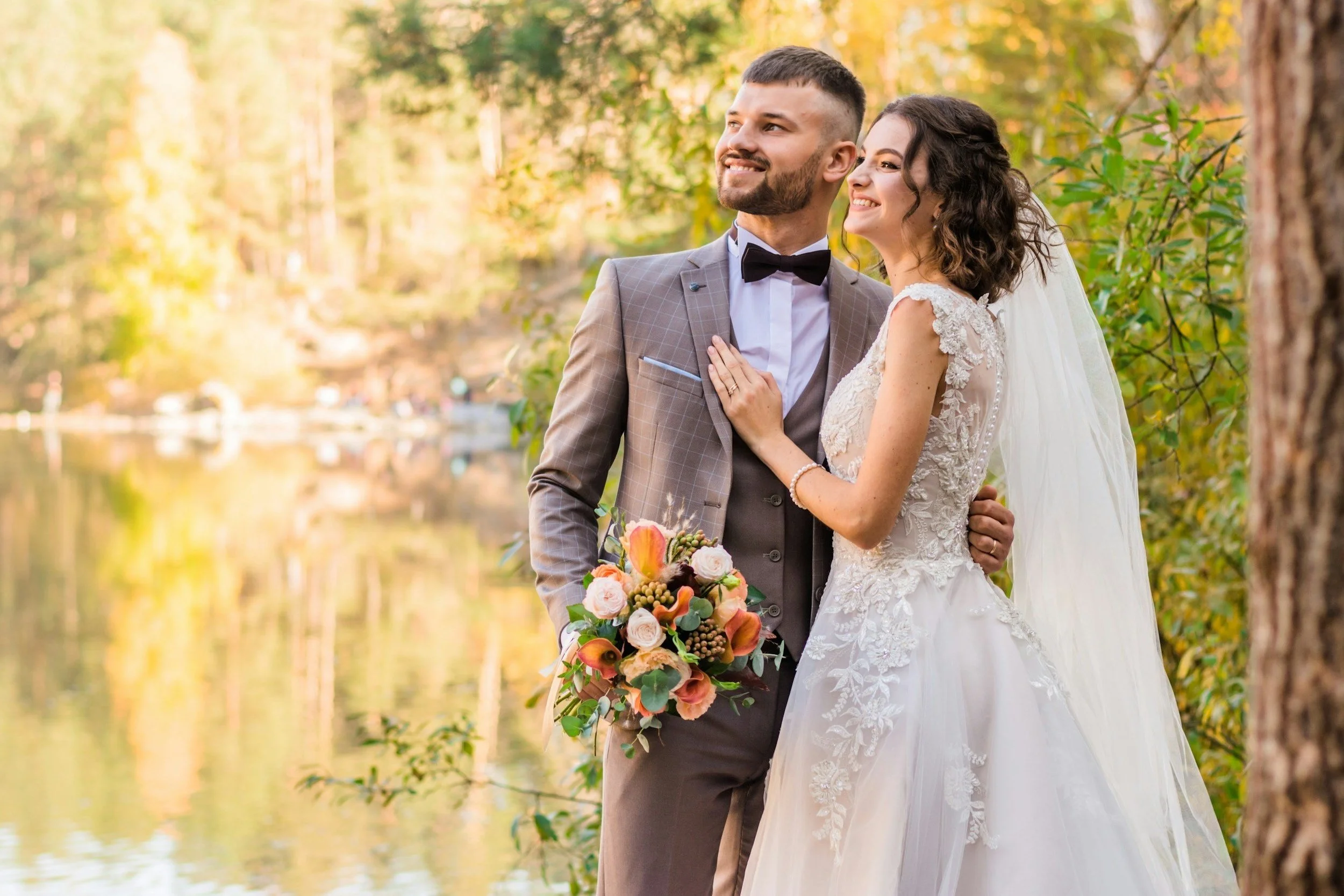 A happy couple in wedding attire standing outdoors near a lake, with a background of trees and autumn foliage.