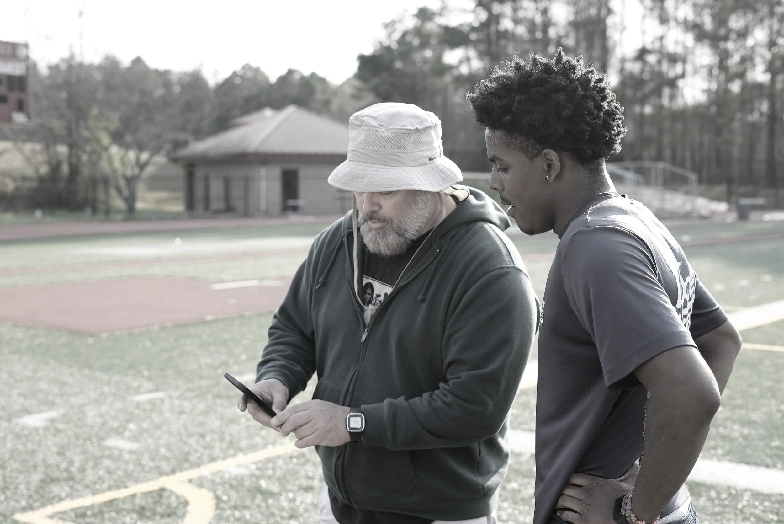 Two men, one with a beard wearing a baseball cap and hoodie, the other with short curly hair, are standing outdoors on a sports field looking at a tablet device.